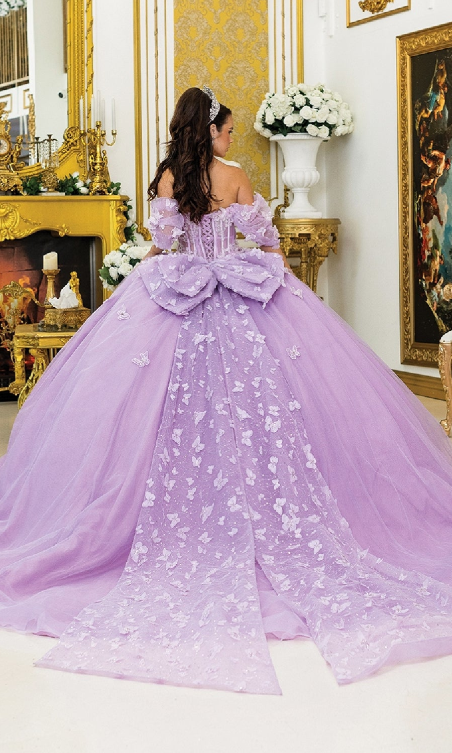 A woman wearing the Purple Quinceanera Dress 5008 by Dancing Queen, featuring butterfly appliques and a large bow, stands in an ornate room decorated with gold accents and white flowers.