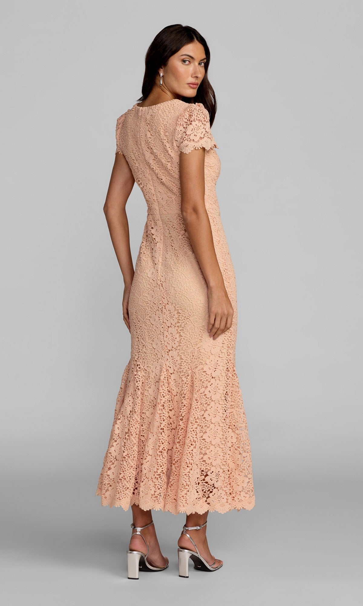 A woman stands facing away against a plain gray background, wearing the Mac Duggal 50116 Tea-Length Party Dress in peach lace with short sleeves, paired with silver high-heeled sandals.