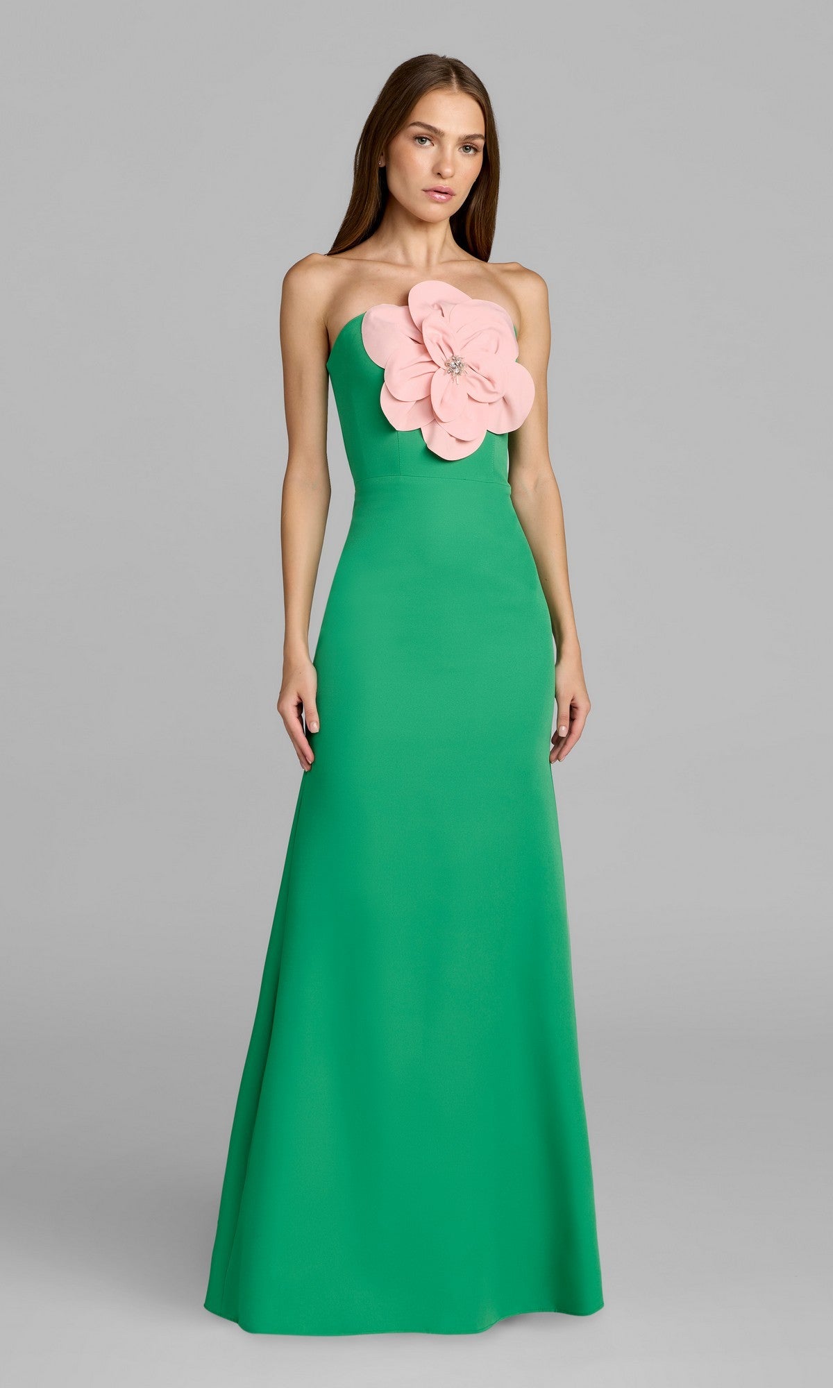A woman stands against a plain gray background wearing the Mac Duggal 50122 long formal dress in spring green, featuring a strapless design with a striking large pink flower embellishment on the chest.