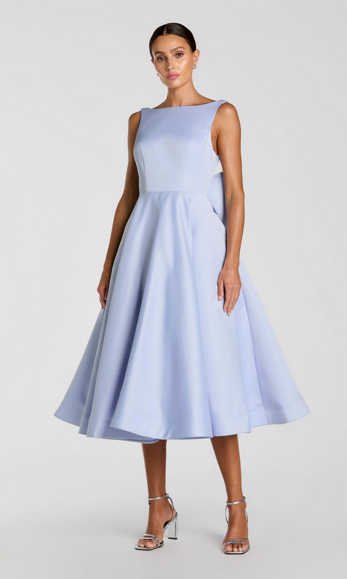 A woman wearing the Tea-Length Party Dress: Mac Duggal 50127 in periwinkle, a sleeveless, knee-length A-line design, paired with silver high-heeled sandals, stands against a plain light background.