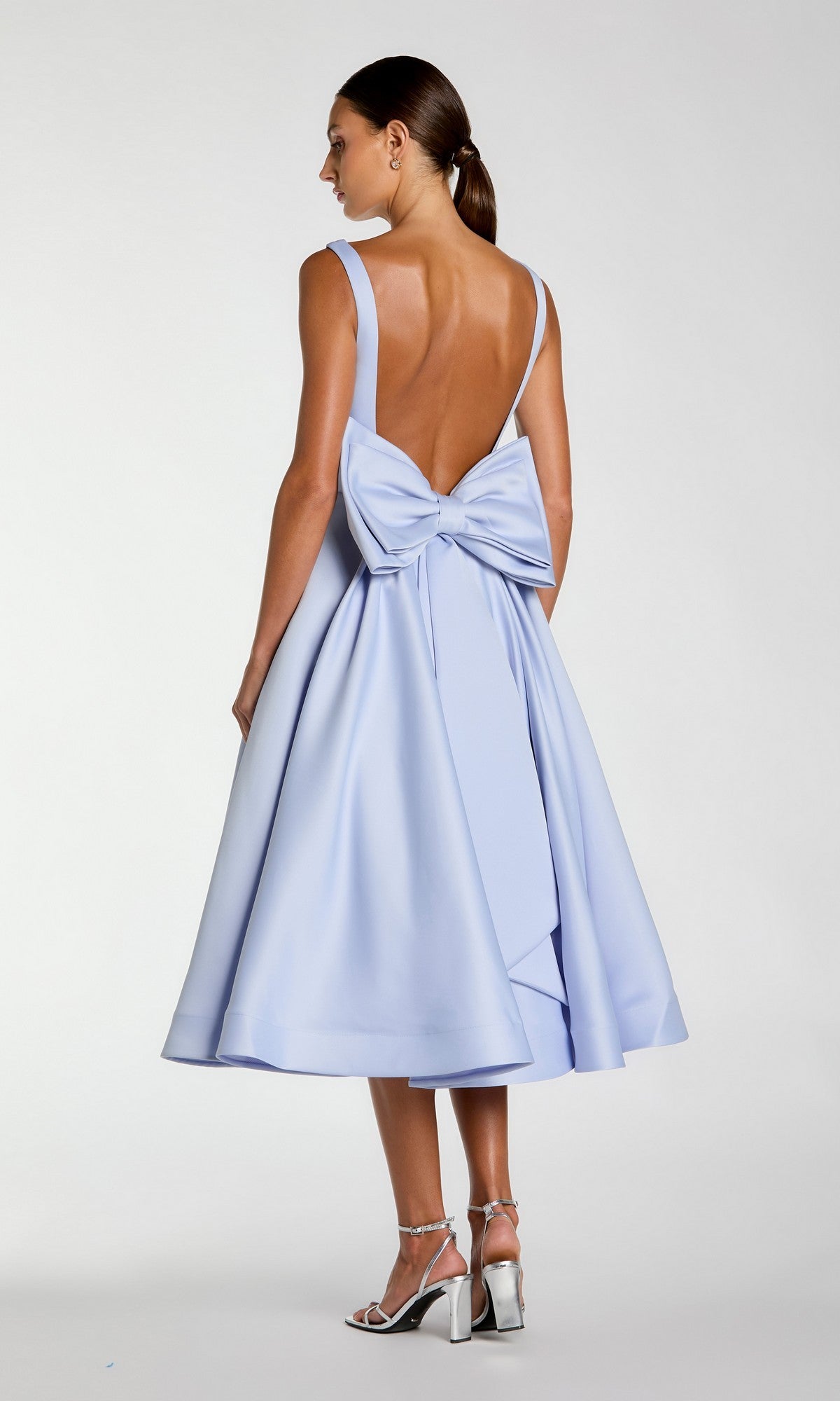 A woman stands against a plain background, wearing the Mac Duggal 50127 Tea-Length Party Dress—a light blue satin design that is sleeveless and backless with a statement bow at the lower back—styled with silver heels and her hair in a low ponytail.