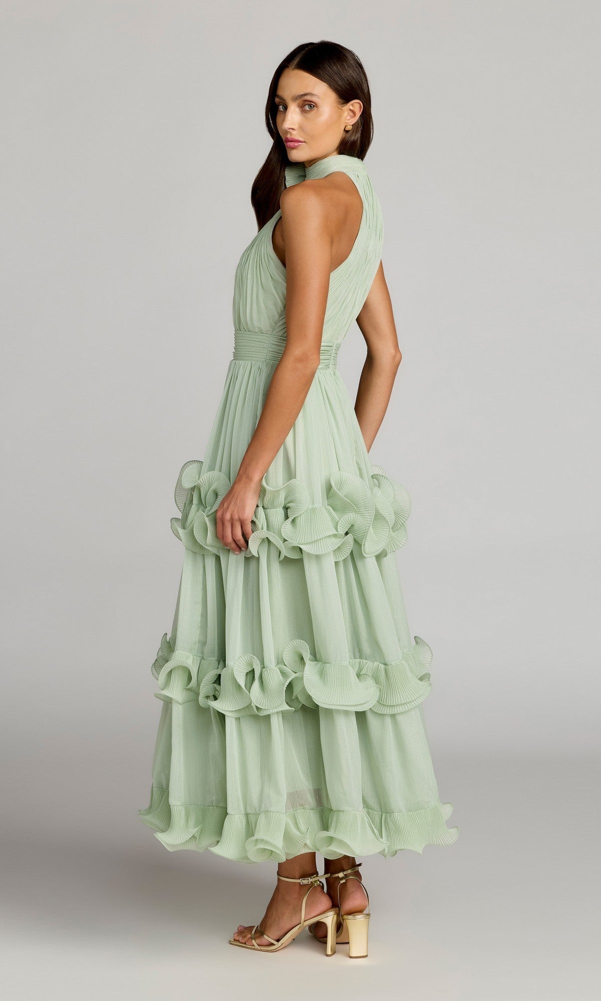 A woman stands sideways against a plain background, wearing the Mac Duggal 50145 long formal dress—a sleeveless, light green ruffled gown with a tiered skirt—paired with gold strappy heels.