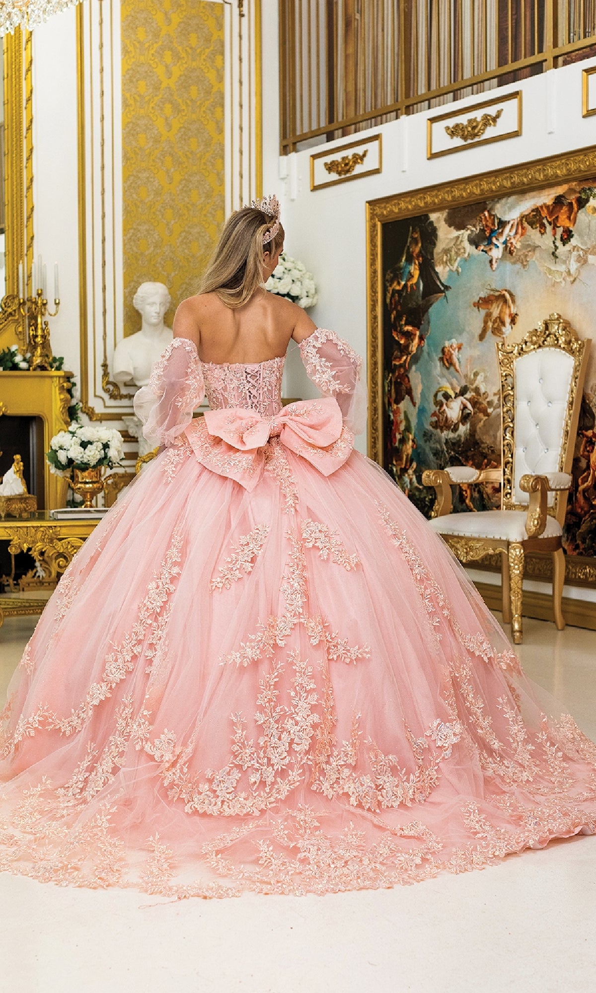 In an ornate room, a woman stands with her back to the camera wearing Quinceanera Dress 5020 By Dancing Queen, a blush pink gown featuring lace details, a corset bodice, and a large bow on the back.