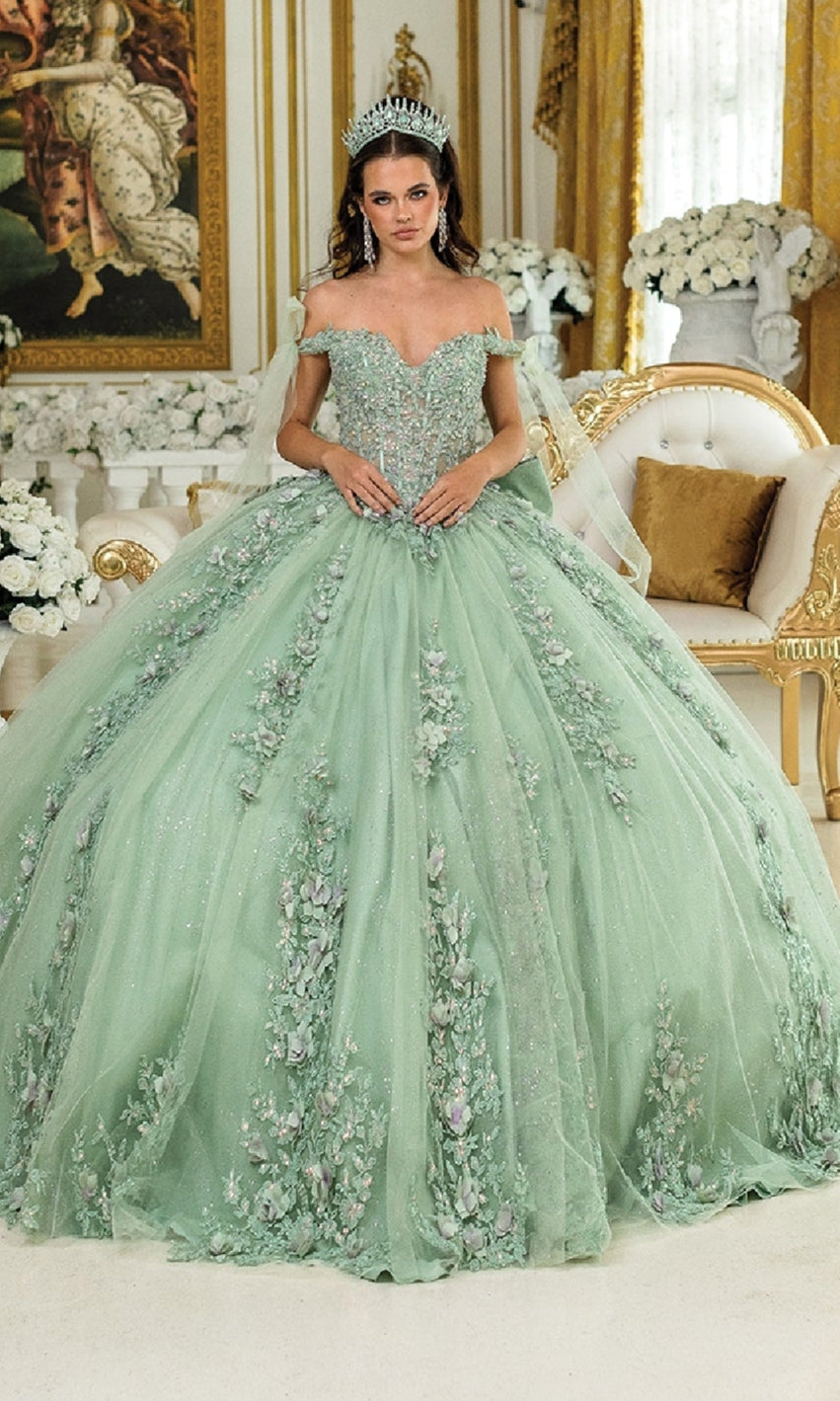 A woman in the Green Quinceanera Dress 5021 By Dancing Queen, featuring sage green fabric and floral embellishments, wears a tiara and stands in an ornate room with gold accents and white flowers.