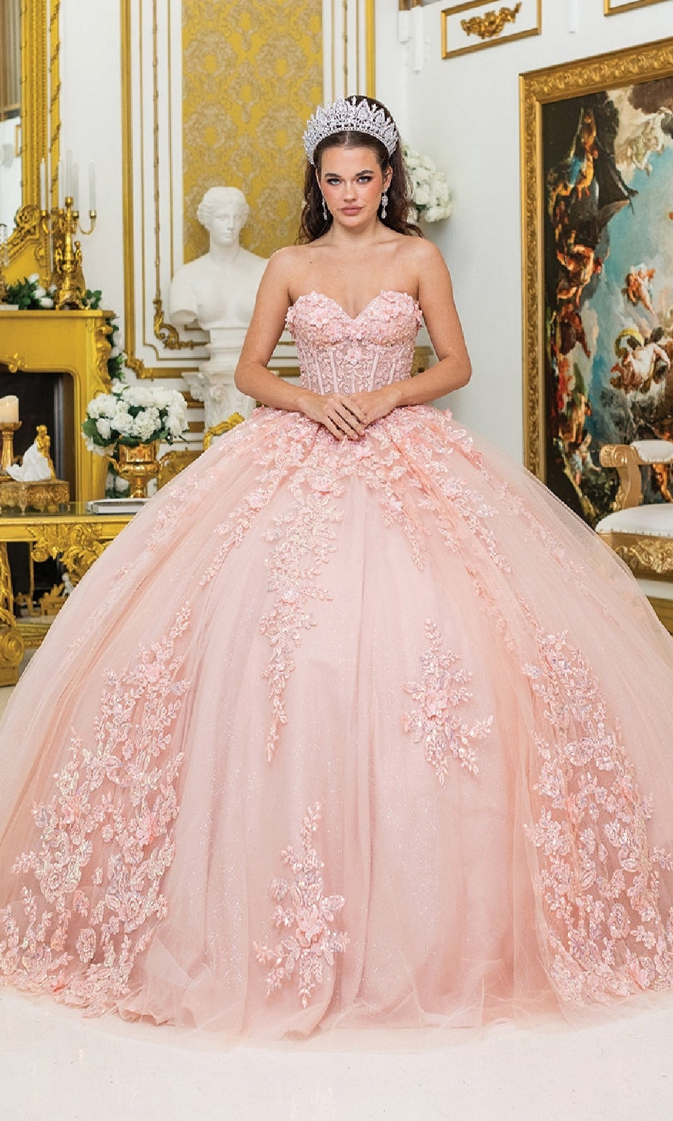 A woman stands in an ornate, gold-accented room filled with classical artwork and statues, wearing the Quinceanera Dress 5027 By Dancing Queen—a sparkling tiara crowns her blush pink ball gown with a corset bodice.