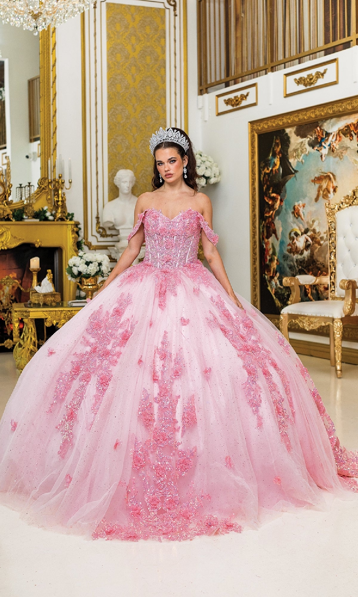 A young woman in the Pink Quinceanera Dress 5034 By Dancing Queen and a sparkling tiara stands in an ornate room with gold accents, a fireplace, and a classical statue in the background.