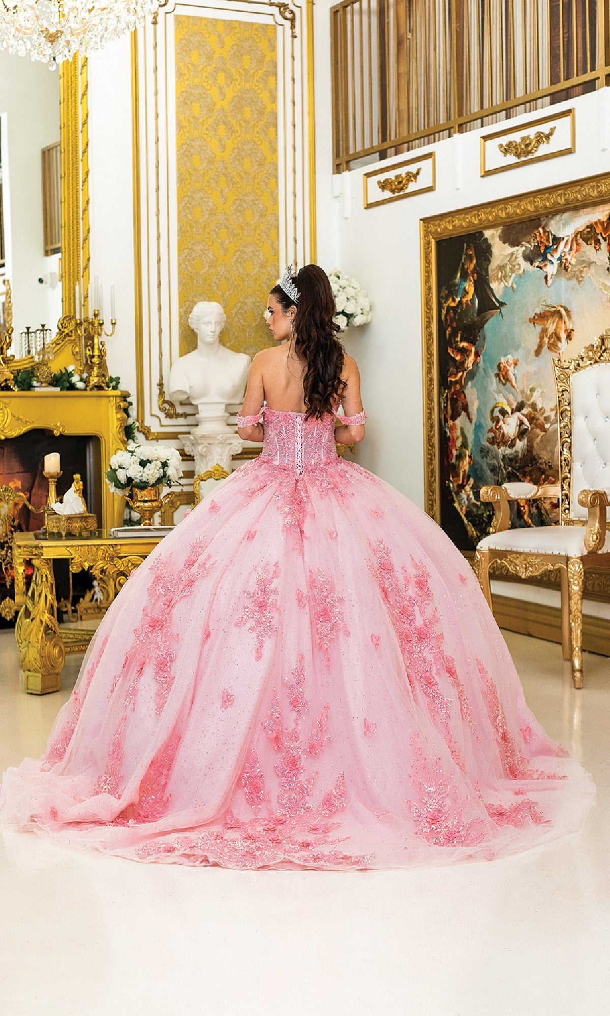 A young woman in the Pink Quinceanera Dress 5034 by Dancing Queen and a tiara stands in an ornate room with gold accents, a fireplace, and classical art, facing away from the camera.