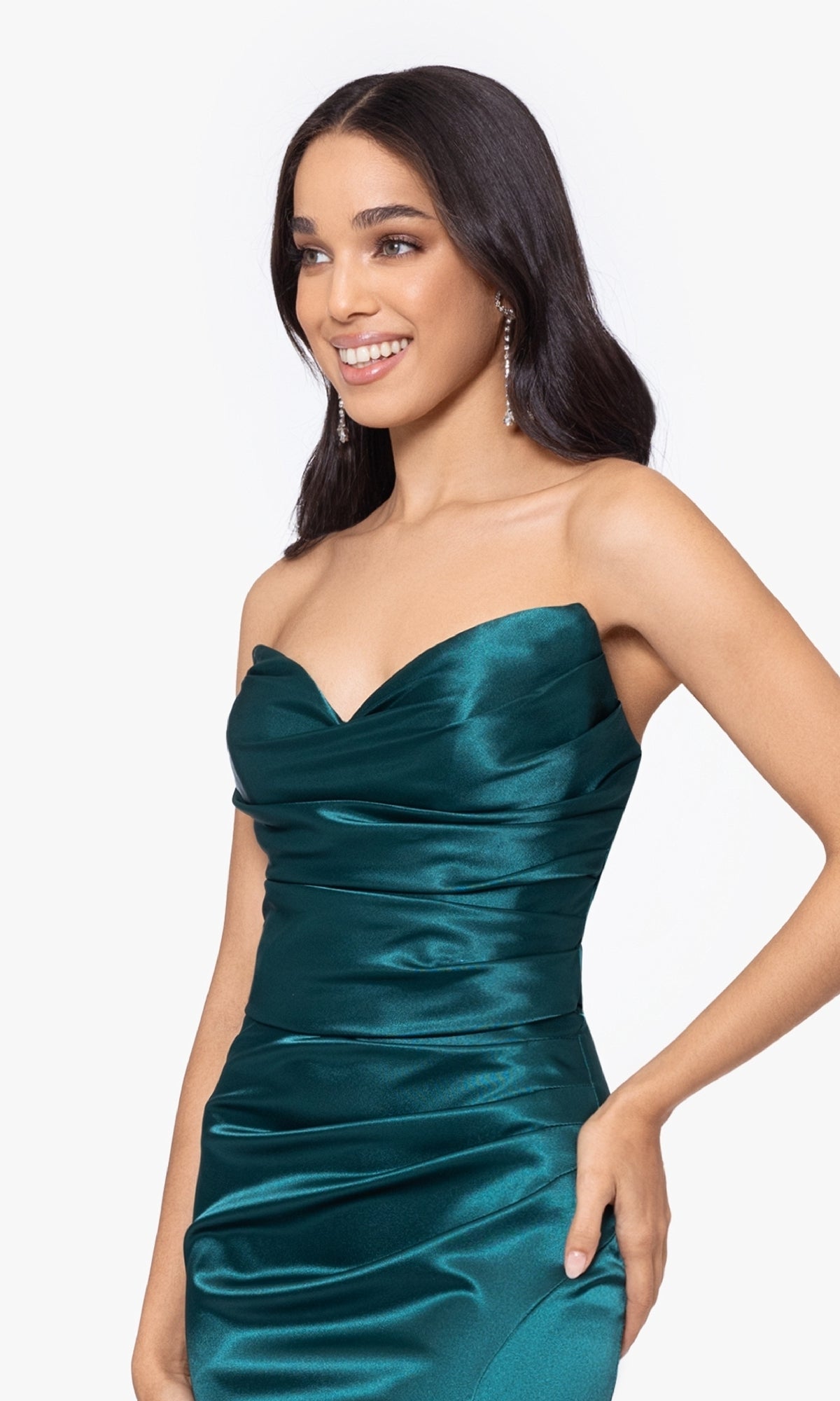 A woman with long dark hair and drop earrings smiles in the Blondie Nites 5041BN strapless long prom dress in emerald green satin, featuring a sweetheart neckline, posing against a plain background.