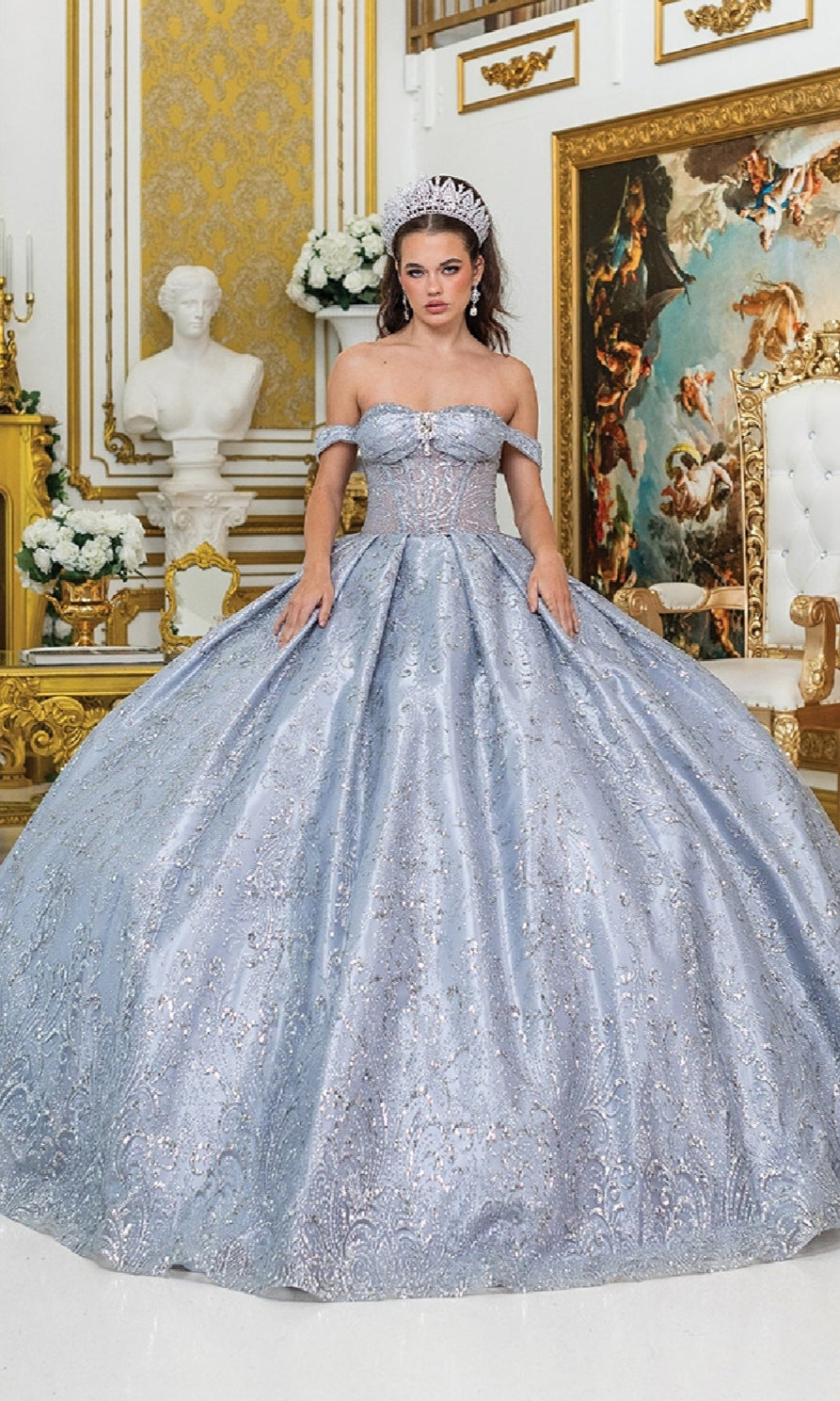 A woman wearing the Quinceanera Dress 5043 by Dancing Queen—a blue tulle ballgown with a corset bodice and a tiara—stands in an ornate room adorned with gold accents, white flowers, a white statue, and a large painting in the background.