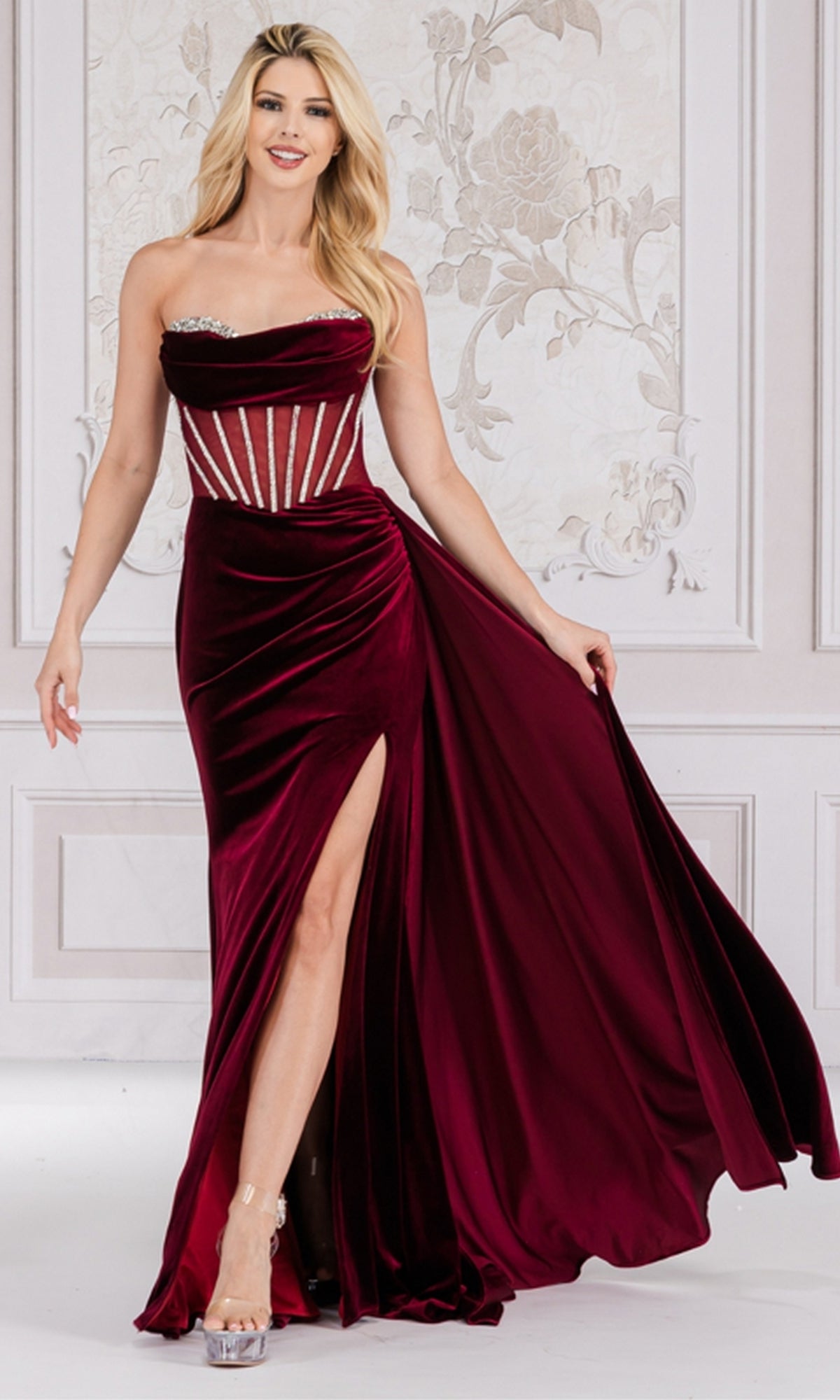 A woman stands in an elegant, light-filled room wearing the Sheer-Waist Strapless Long Velvet Prom Dress 5051, featuring a beaded sheer corset bodice, high slit, and silver accents.