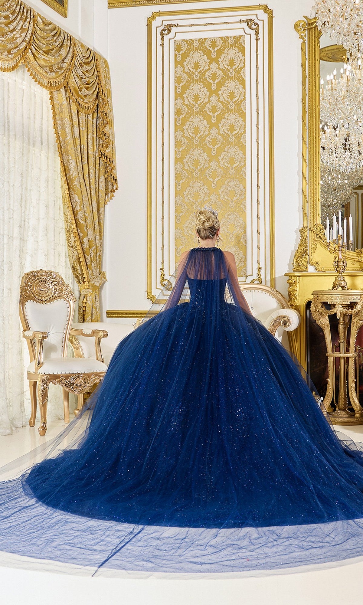 Wearing the Quinceanera Dress 5061 By Dancing Queen, a person stands facing an ornate gold-accented wall in an opulent room filled with gilded furniture and chandeliers. The dark blue gown features glitter tulle and beaded applique.