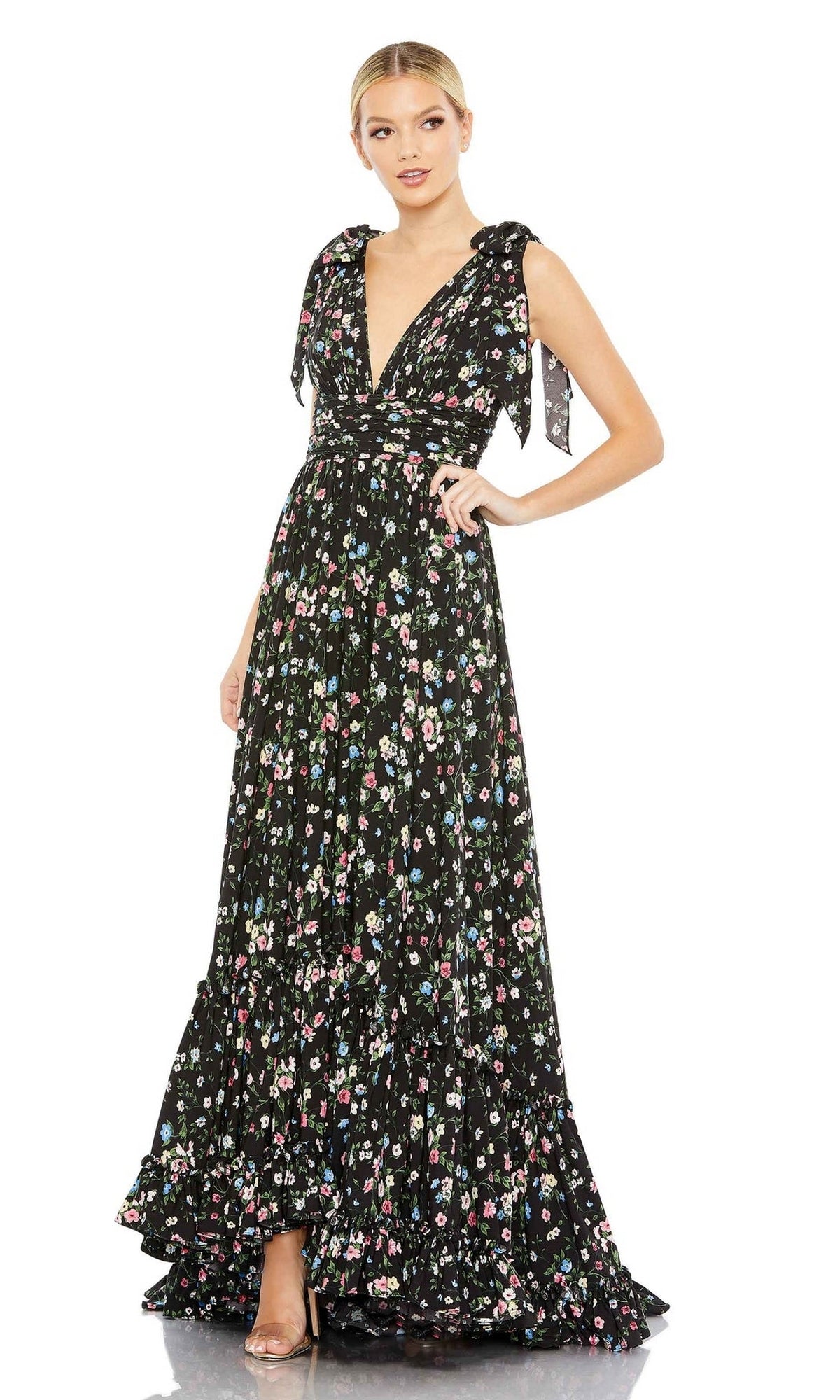 A person confidently poses with one hand on their hip, wearing the Long Formal Dress 50657 by Mac Duggal—a sleeveless, black floral print gown with a deep V-neckline.