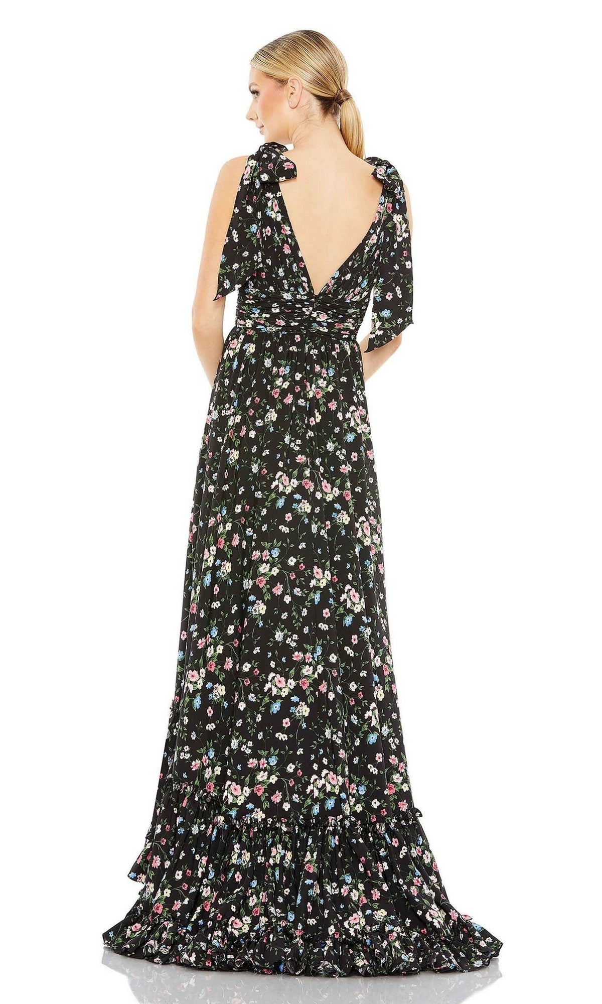A woman stands facing away from the camera, wearing the Long Formal Dress 50657 by Mac Duggal—a long black floral print gown featuring an open back and shoulder ties.
