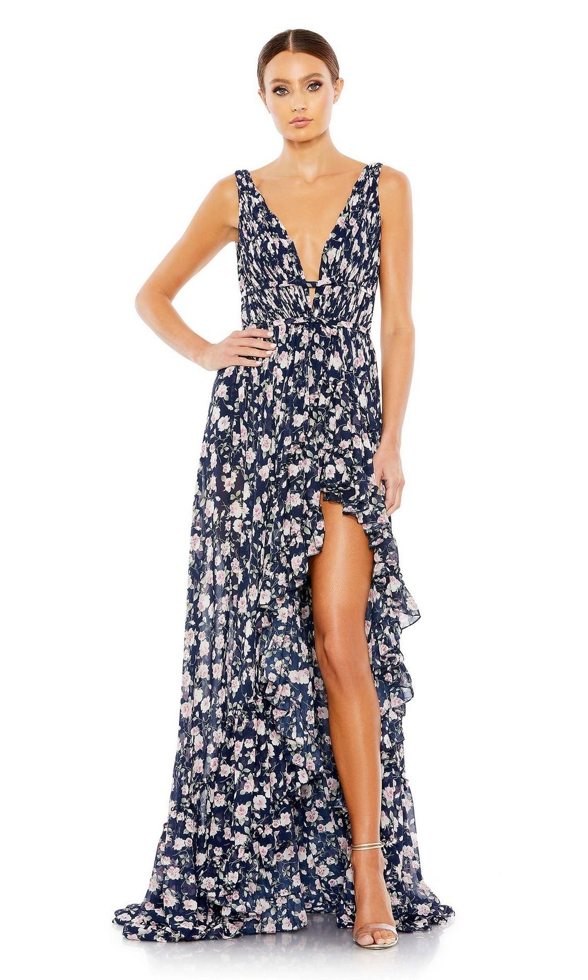 A woman models the Mac Duggal 50659 Floral-Print Maxi Party Dress—sleeveless, deep V-neck, navy blue with a ruffled high slit—paired with strappy heels, standing against a plain white background.