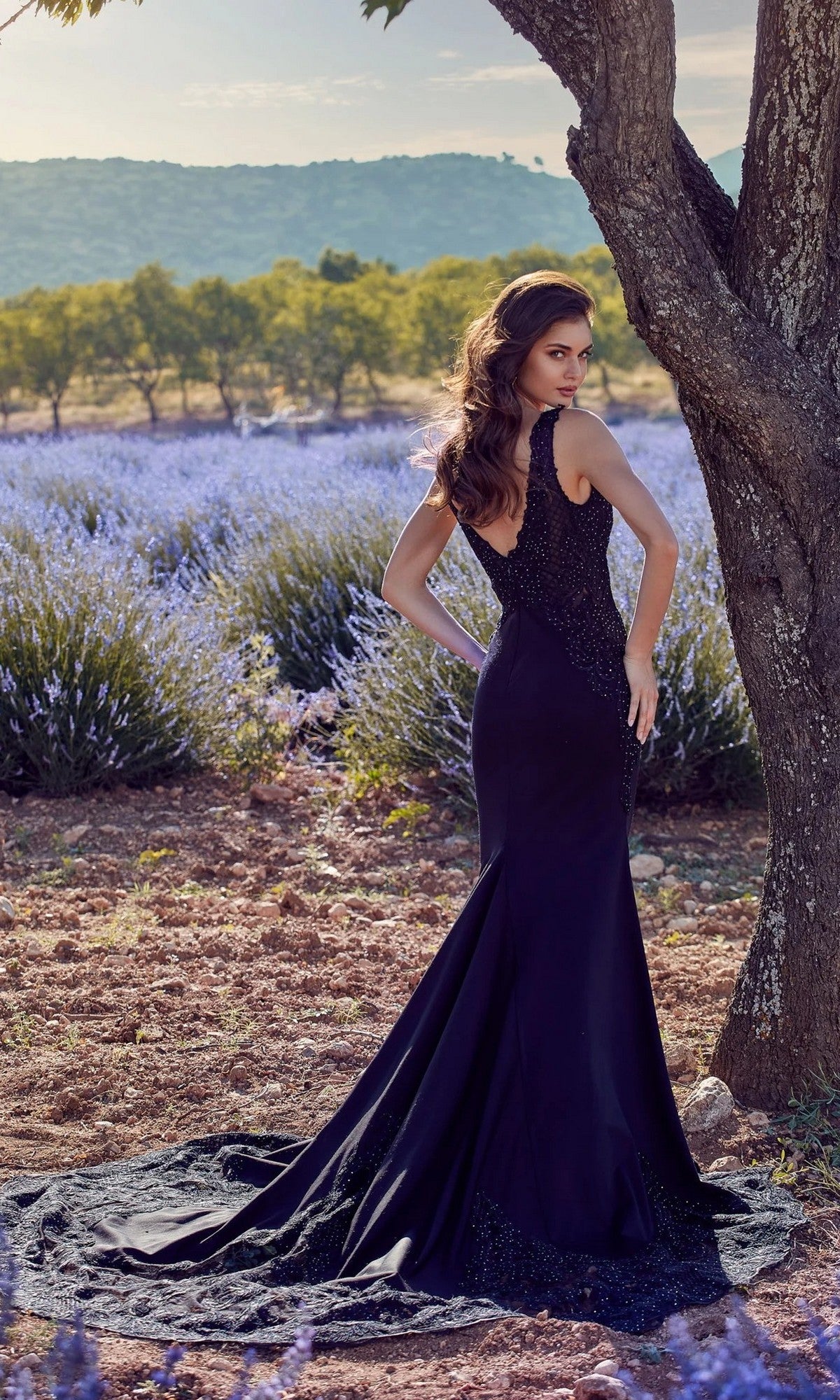 A woman wearing the Alyce 5065 long formal dress stands outdoors on a dirt path near a tree, with lavender bushes and hills in the background.