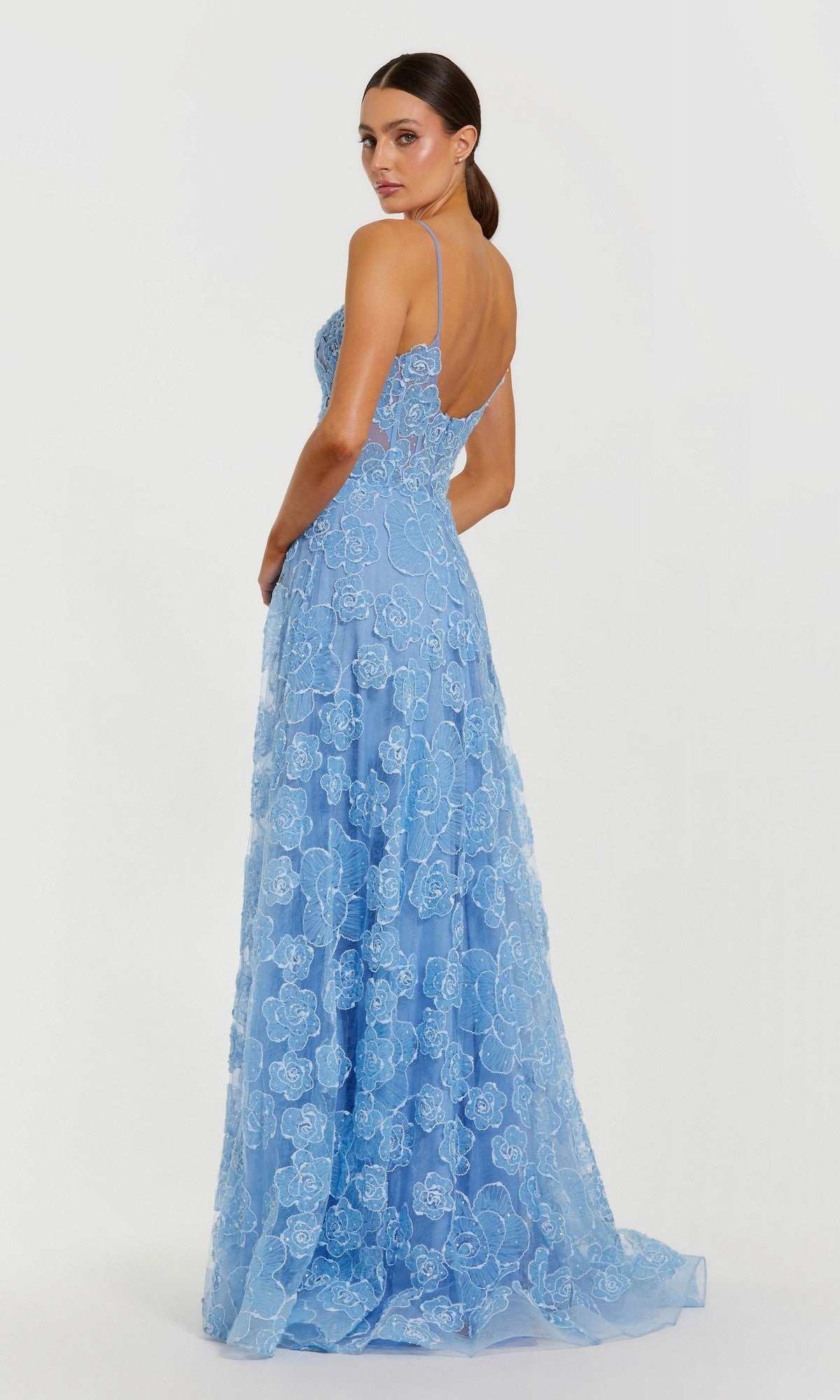A woman models the Mac Duggal 50735 long formal dress in light blue, featuring floral lace details, thin straps, and an elegant floor-length silhouette as she stands with her back slightly turned.