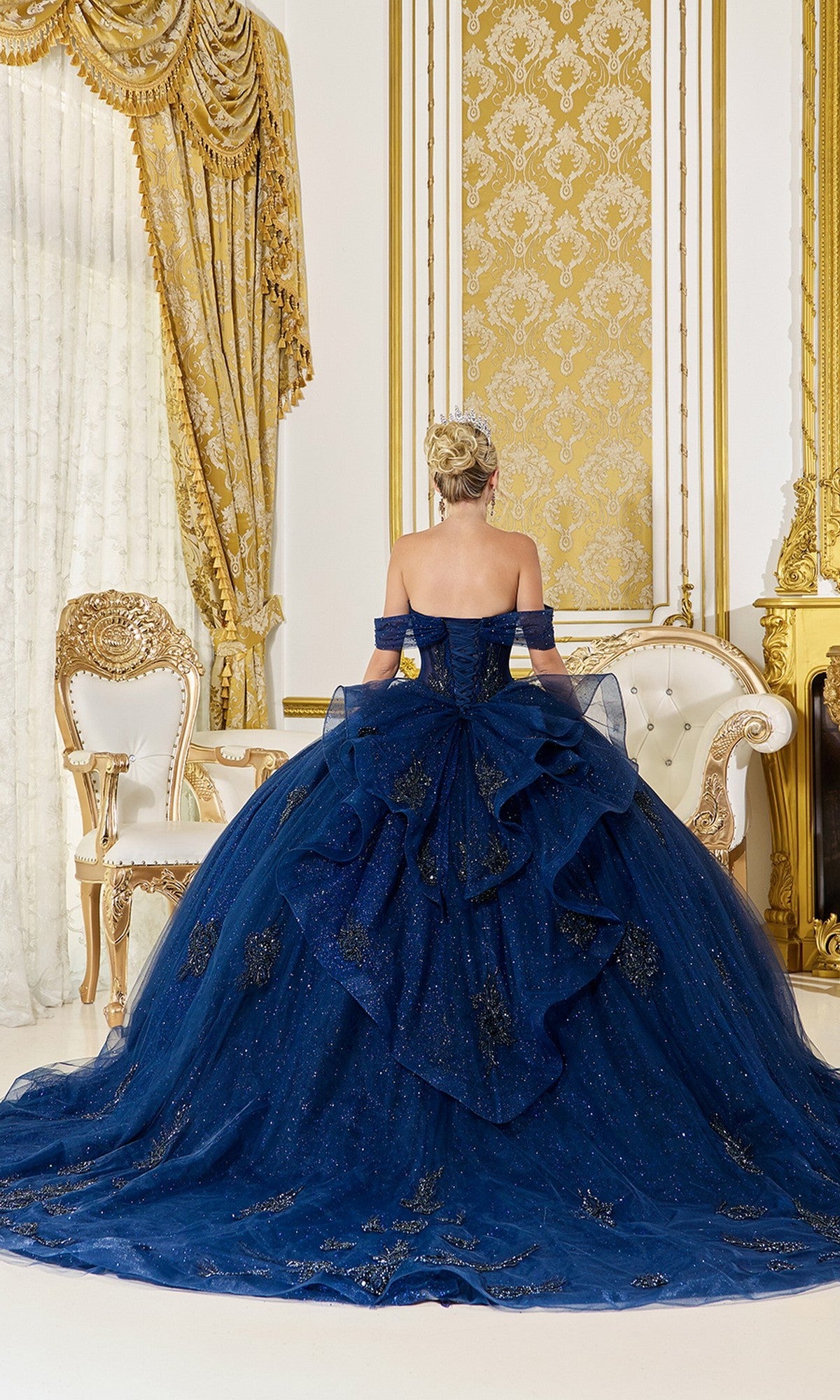 A person in the Quinceanera Dress 5075 By Dancing Queen, an off-the-shoulder navy blue ball gown with gold details, stands with their back to the camera in an ornate room featuring elegant white and gold furniture.