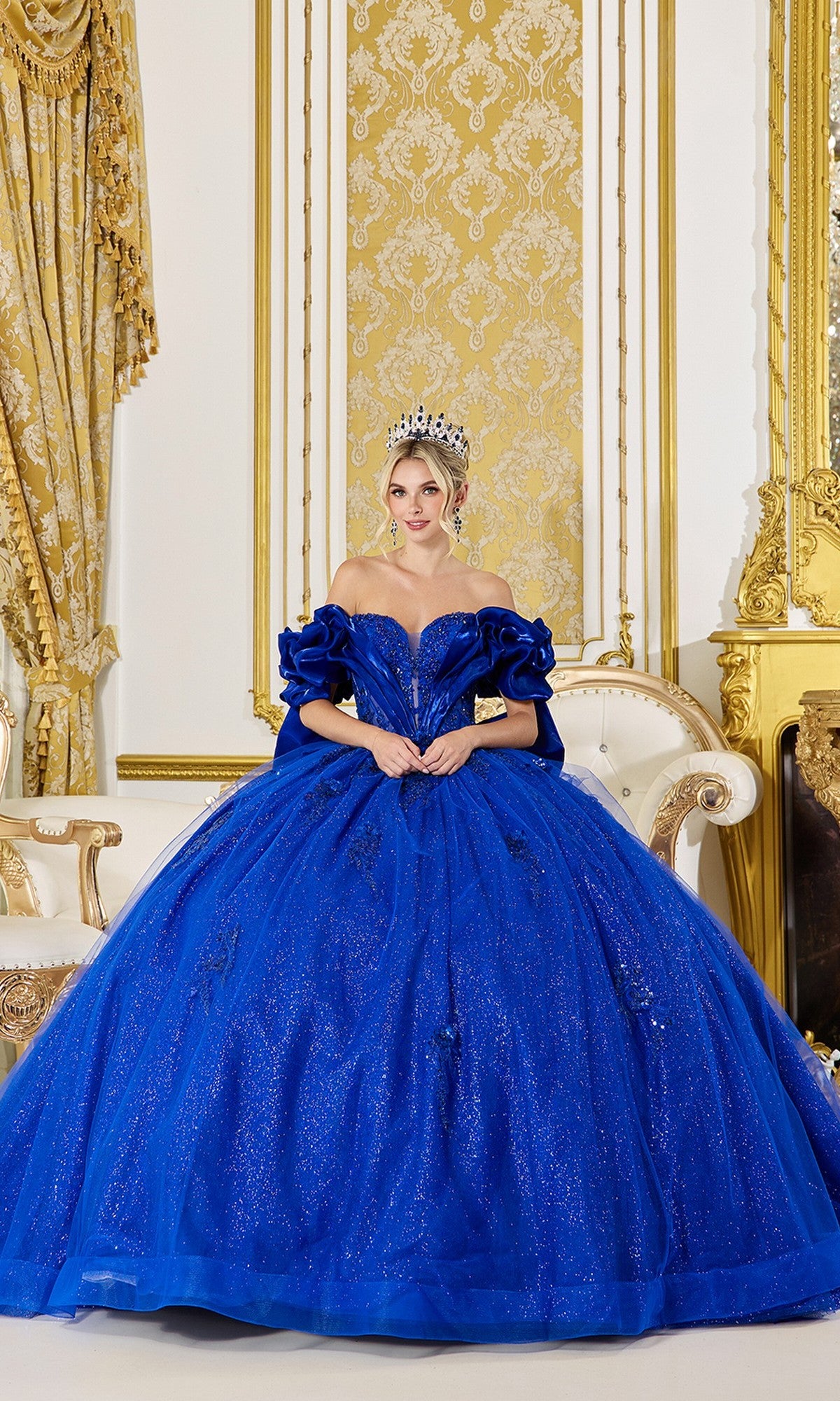 Wearing the Quinceanera Dress 5080 By Dancing Queen in royal blue, a woman with a crown sits on an ornate white and gold sofa in an opulent gold room.