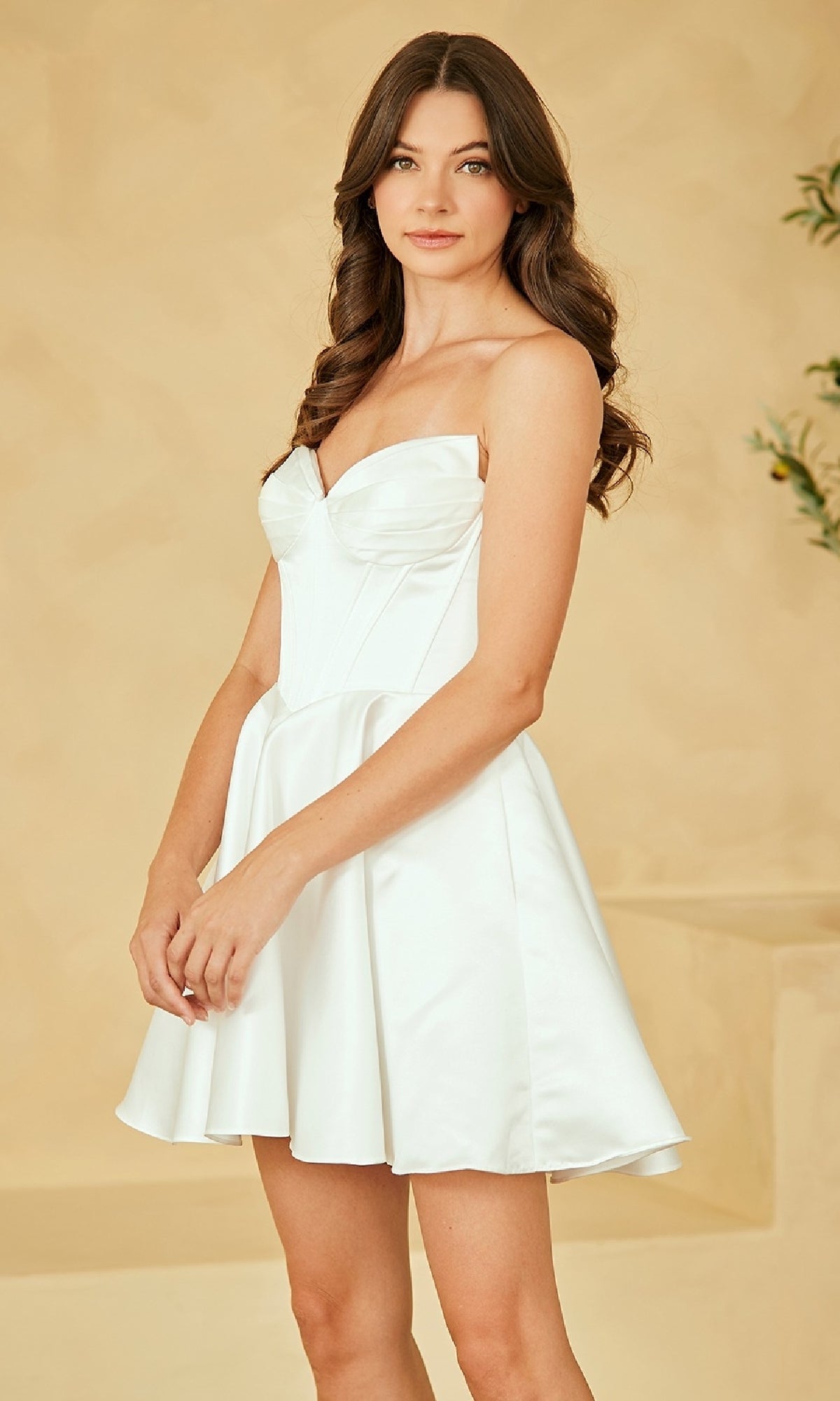 A woman with long brown hair wears the Amelia Couture 5082S strapless white party dress and stands against a beige background.