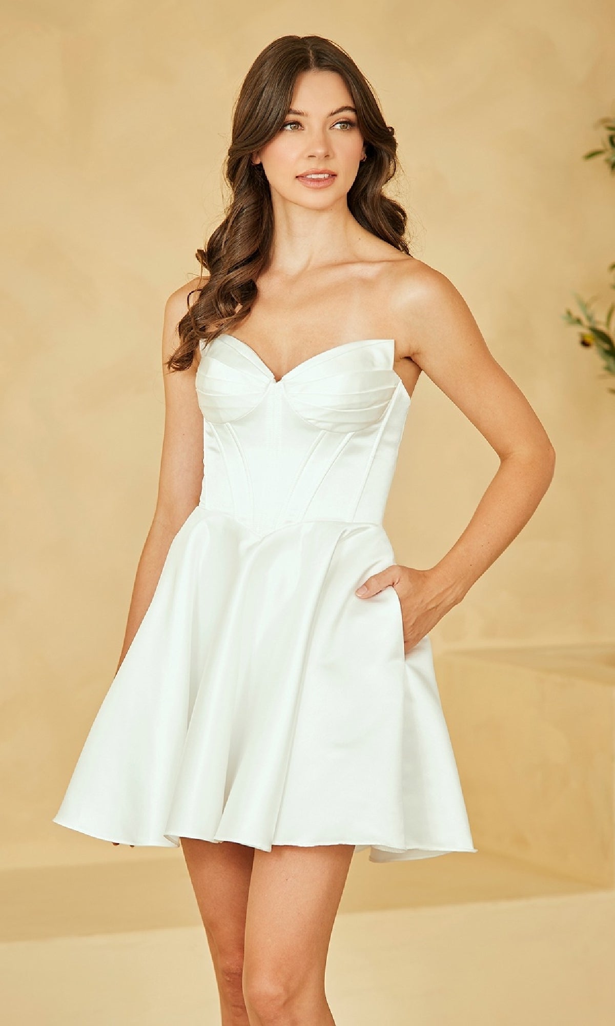 A woman models the Amelia Couture 5082S Strapless White Party Dress, featuring a fitted bodice and flared satin skirt, against a beige background.