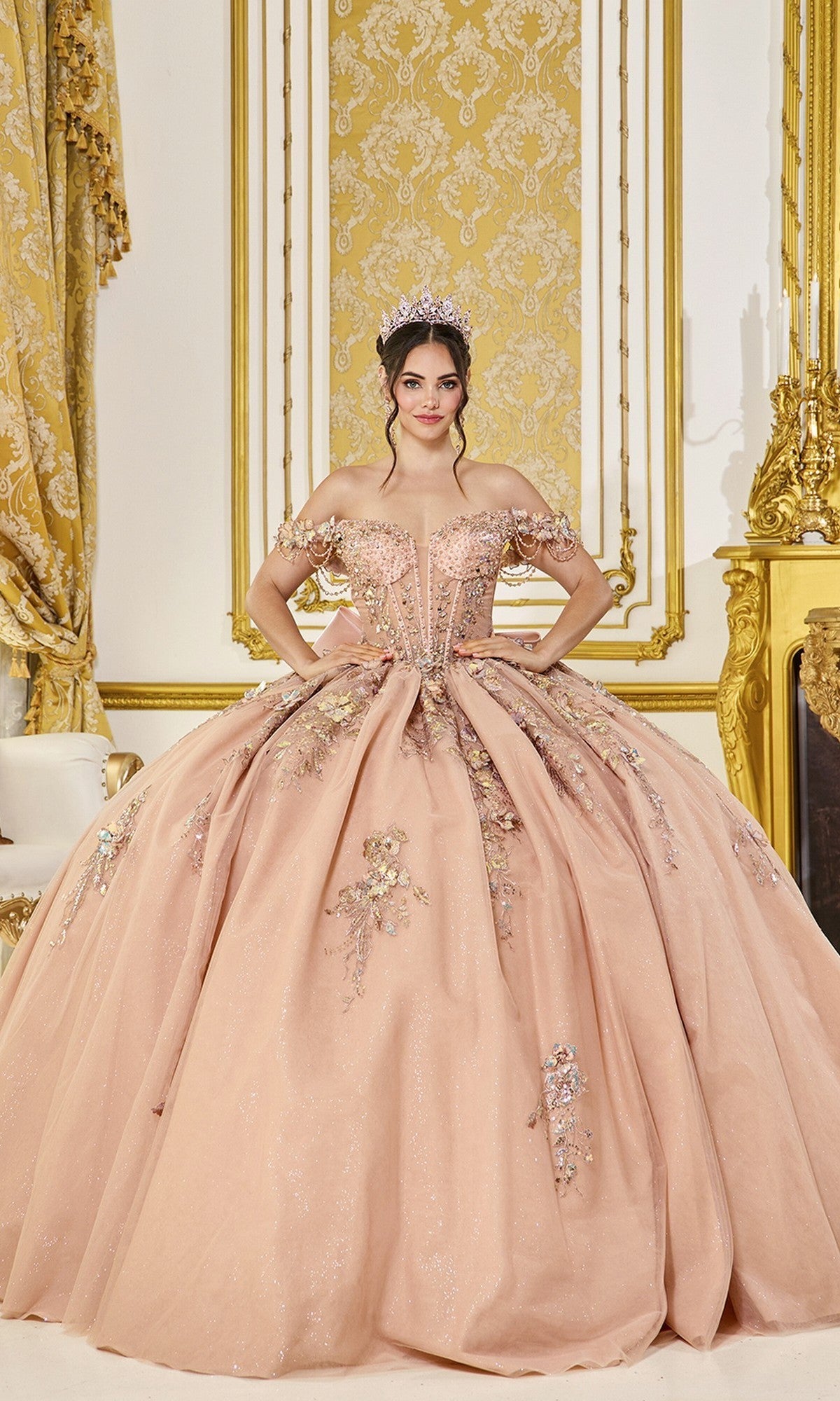 A woman wearing the Quinceanera Dress 5097 By Dancing Queen in rose gold, featuring an off-the-shoulder design and embellished corset bodice with a tiara, stands in an ornate room adorned with gold and cream curtains and matching wall paneling.
