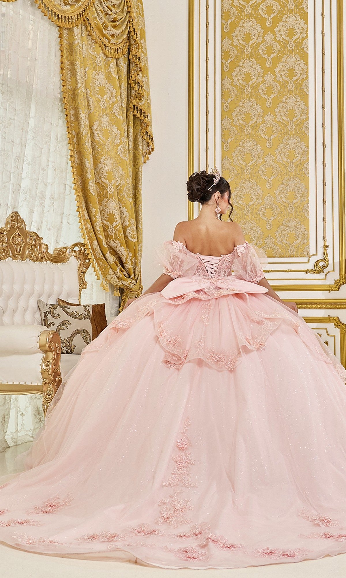 A woman in the Quinceanera Dress 5099 By Dancing Queen, a blush pink ball gown with a corset bodice, stands with her back to the camera in an ornate room with gold-patterned walls, matching curtains, and a white and gold sofa.