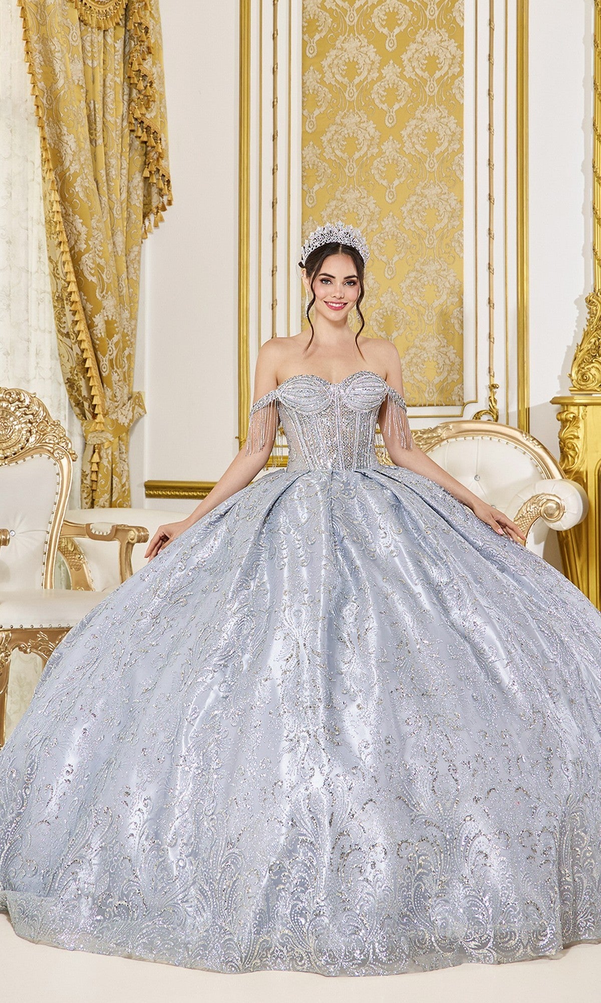 A young woman poses indoors in a gold and white ornate room wearing the Quinceanera Dress 5107 By Dancing Queen in silver, featuring off-the-shoulder sleeves and sparkly embellishments, paired with a tiara.