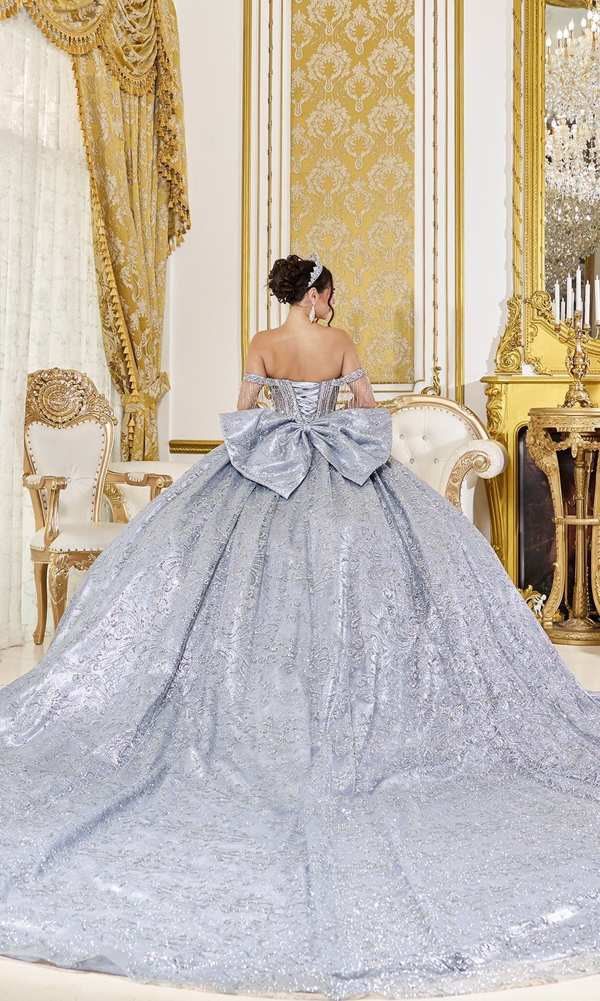 A woman wearing the Quinceanera Dress 5107 by Dancing Queen—an off-the-shoulder silver ball gown with a large bow and sparkly accents—stands in an ornate gold-decorated room, facing away from the camera.