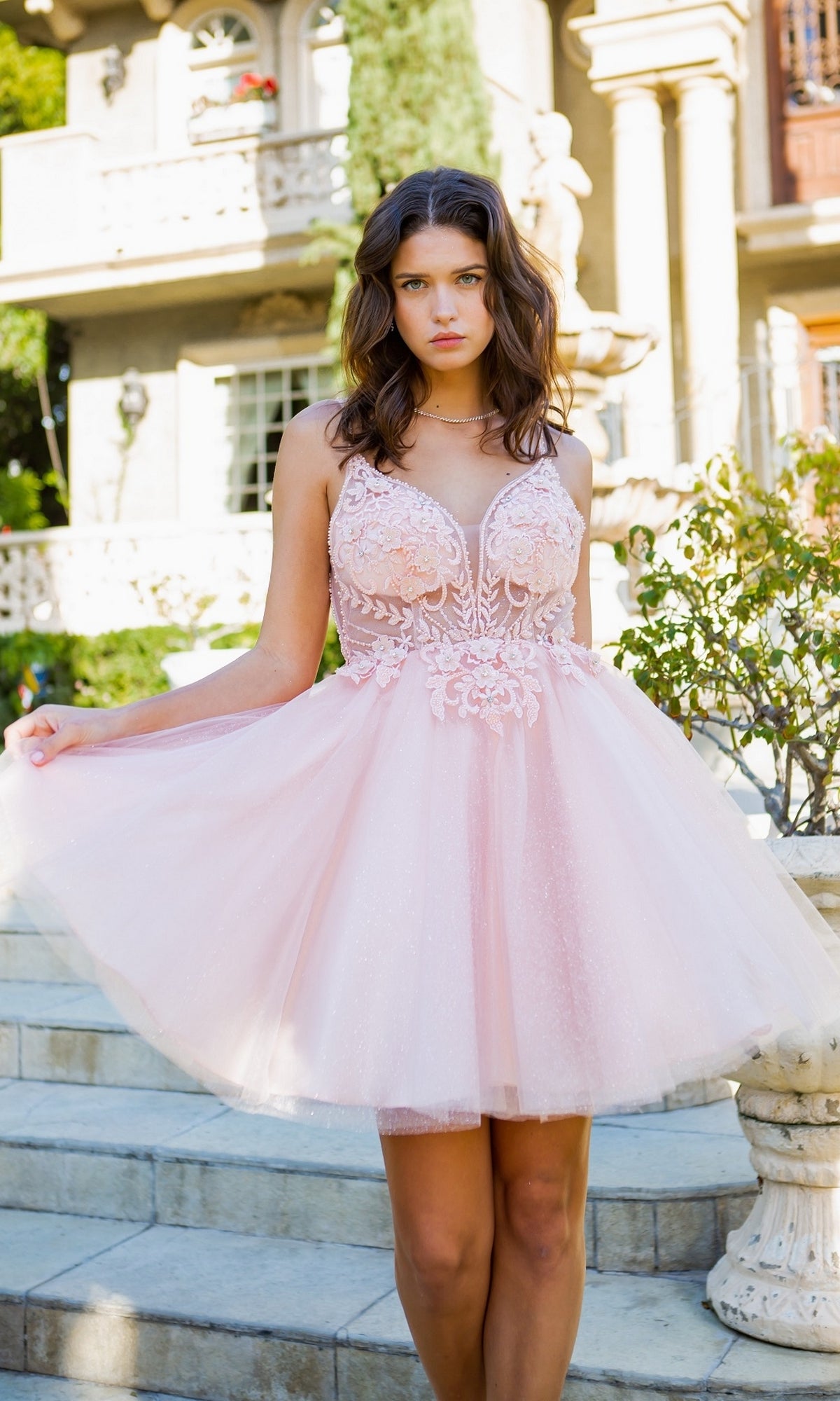 A young woman stands on stone steps outdoors, wearing the Short Ball-Gown-Style Homecoming Dress 5112J in blush pink lace and tulle, with a building and greenery behind her.