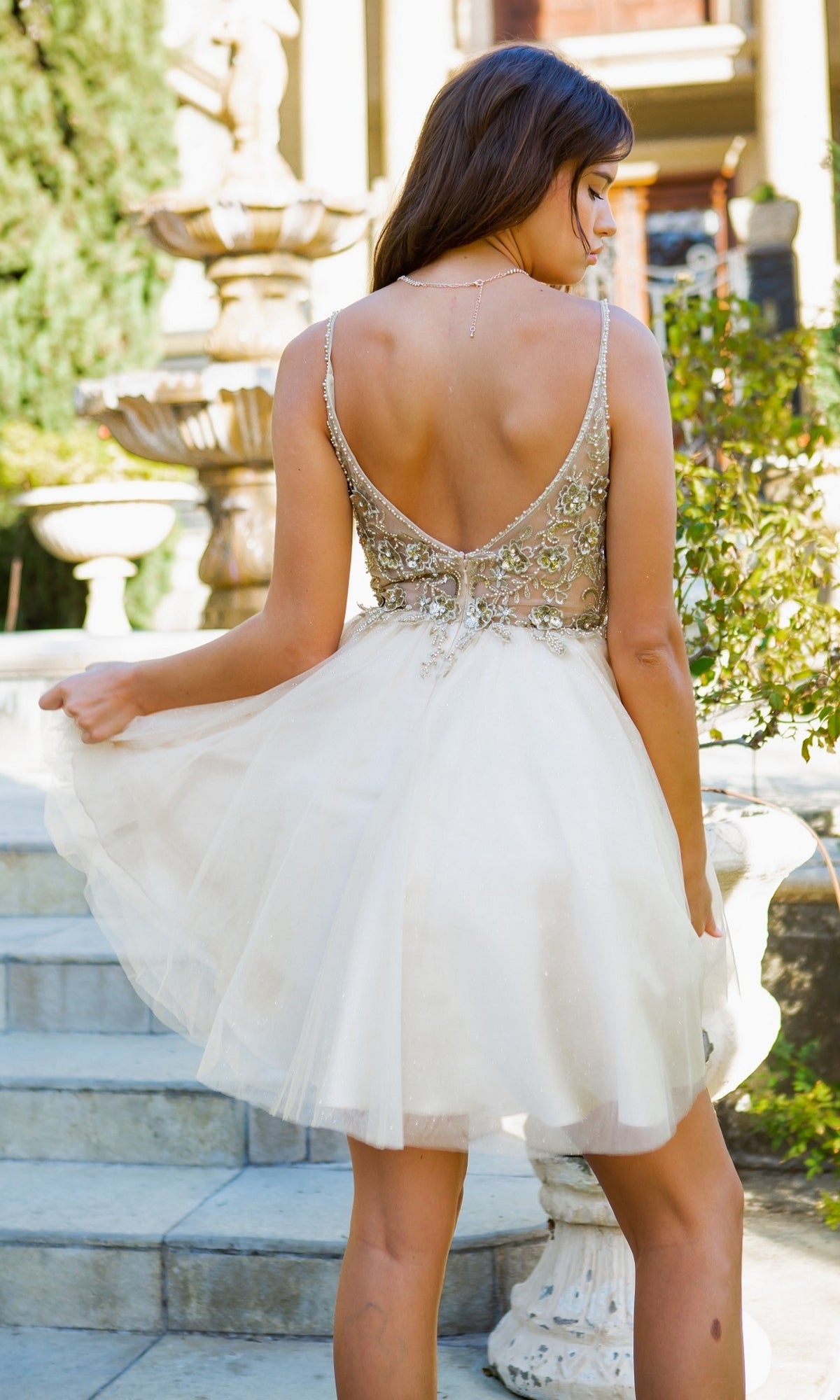 Wearing the Short Ball-Gown-Style Homecoming Dress 5112J—a sleeveless, backless white dress with intricate beadwork—a woman stands outdoors on stone steps by a fountain, holding the skirt and facing away.