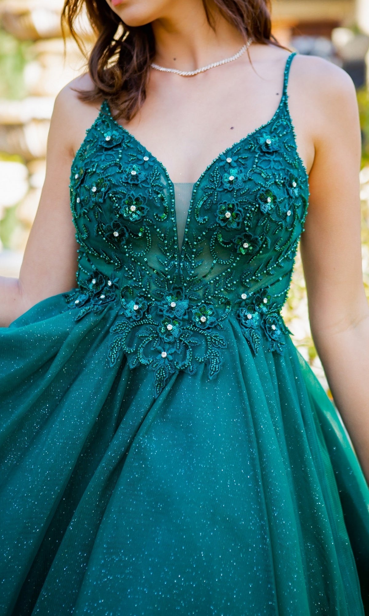A woman wears the Short Ball-Gown-Style Homecoming Dress 5112J, a sleeveless deep green dress with floral embroidery, bead embellishments, a sheer neckline panel, and a full sparkly skirt.