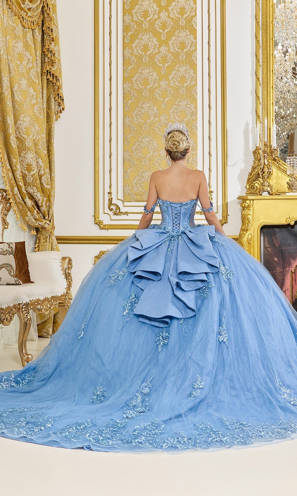 A woman wearing the Quinceanera Dress 5117 by Dancing Queen, featuring a glittering tulle ballgown and corset bodice, stands with her back to the camera in an ornate room adorned with gold accents.