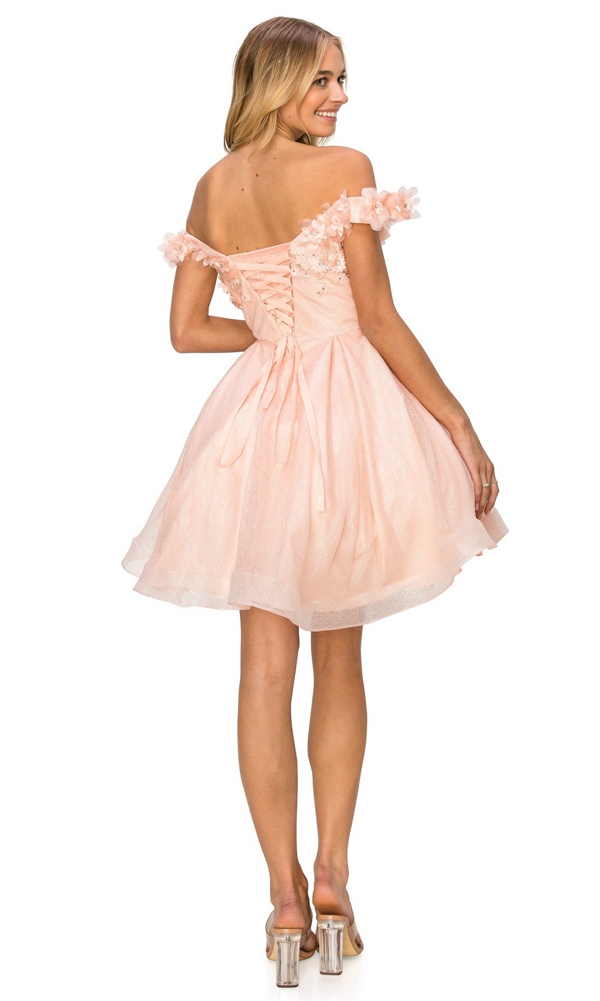 Woman in the Shimmer Off-Shoulder Short Homecoming Dress 5120J, a light pink dress with floral accents and a lace-up corset back, stands facing away in clear heeled shoes.