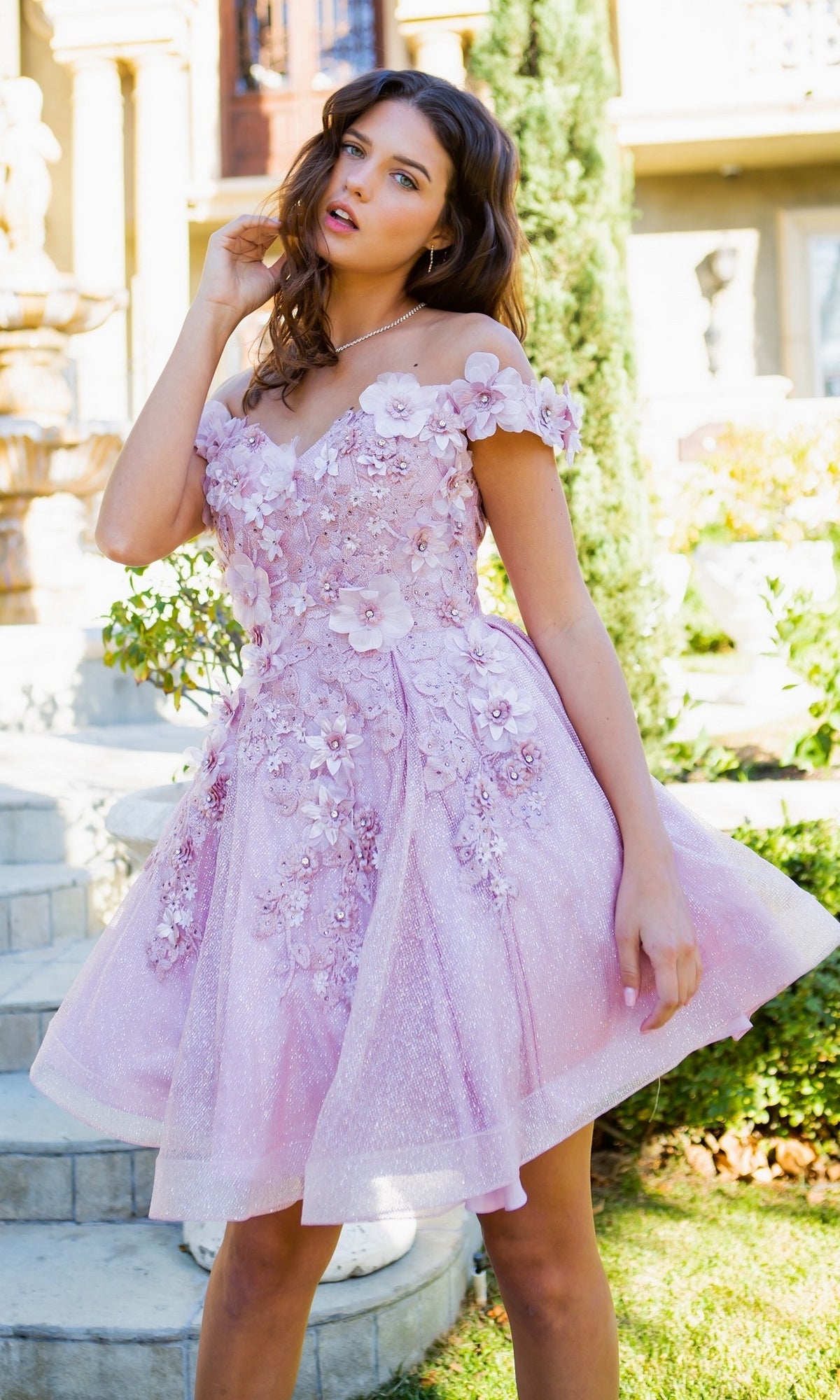 A woman poses outdoors in the Shimmer Off-Shoulder Short Homecoming Dress 5120J—a light pink, off-the-shoulder dress with floral embellishments and a flared baby doll party silhouette.