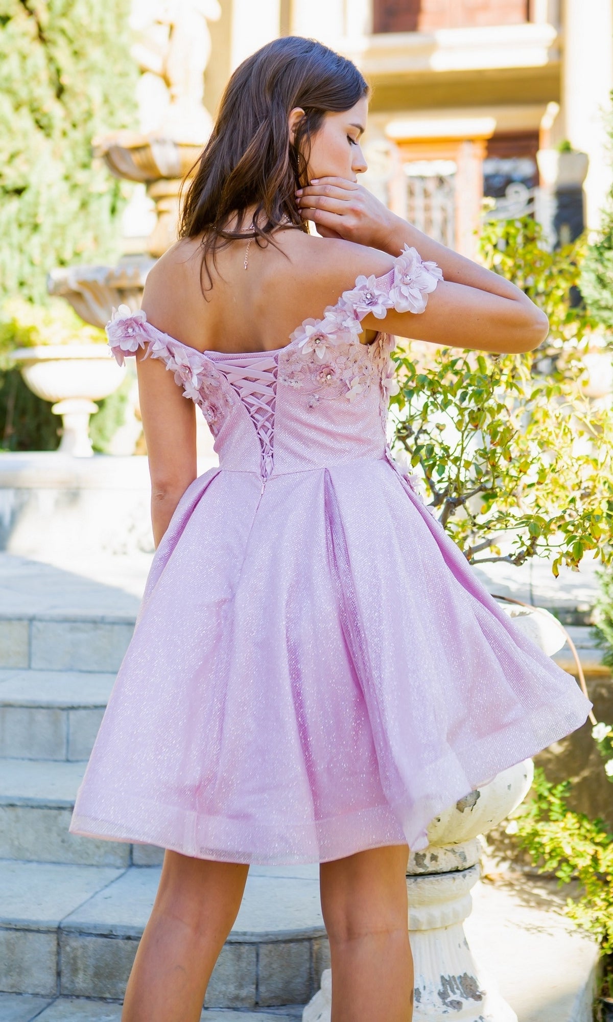 A woman stands on outdoor steps wearing the Shimmer Off-Shoulder Short Homecoming Dress 5120J in light pink with floral details and a corset back, perfect for parties.