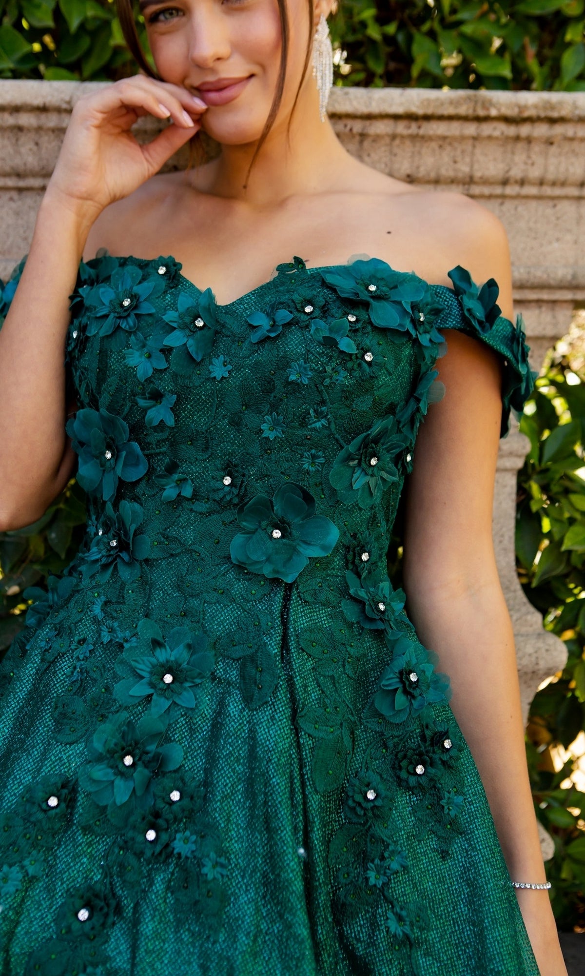 A woman in the Shimmer Off-Shoulder Short Homecoming Dress 5120J, featuring a corset back, emerald green hue, 3D floral appliqué, and pearl accents, stands outdoors beside a stone railing.