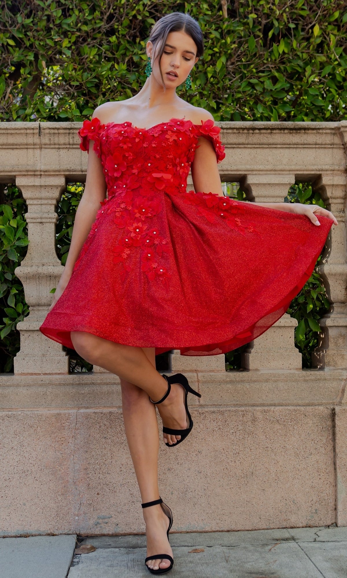 A woman in the Shimmer Off-Shoulder Short Homecoming Dress 5120J, featuring floral embellishments, stands by a stone railing and greenery, gracefully holding out one side of her knee-length skirt.