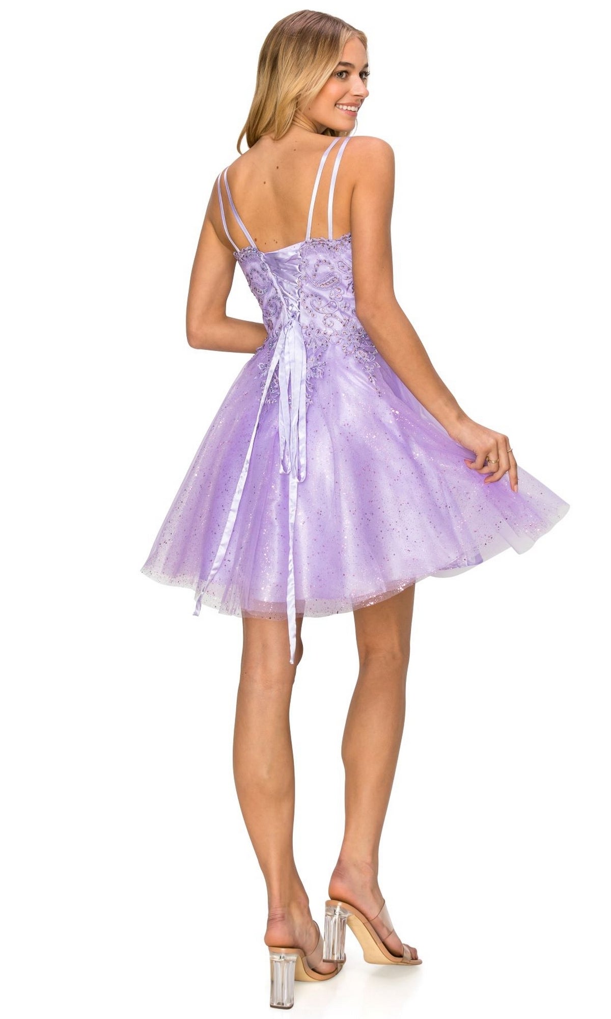 A woman in the Corset-Back Short Glitter Homecoming Dress 5125J, featuring double spaghetti straps and a lace-up back, stands in clear heels facing away and looks over her shoulder.