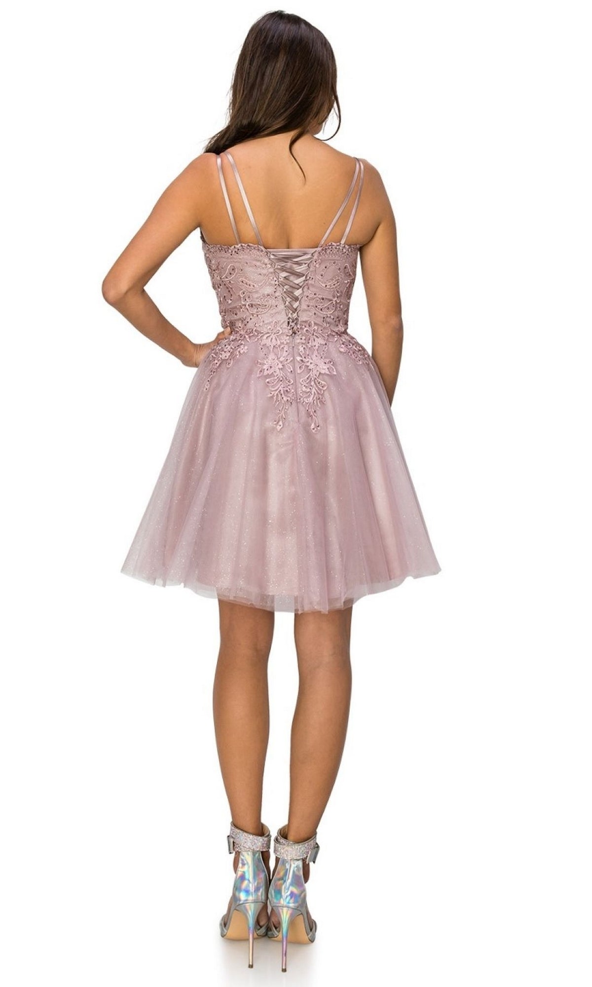 A woman faces away, wearing the Corset-Back Short Glitter Homecoming Dress 5125J with a lace-up back, thin straps, and iridescent high-heeled boots.