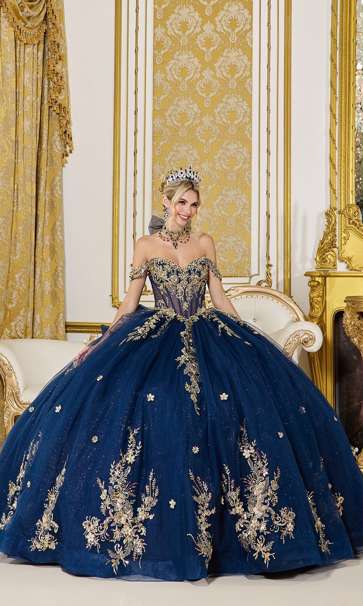 A woman in the Quinceanera Dress 5128 By Dancing Queen, featuring navy fabric with gold applique and a jeweled tiara, sits on a cream sofa in an opulent room with gold accents.