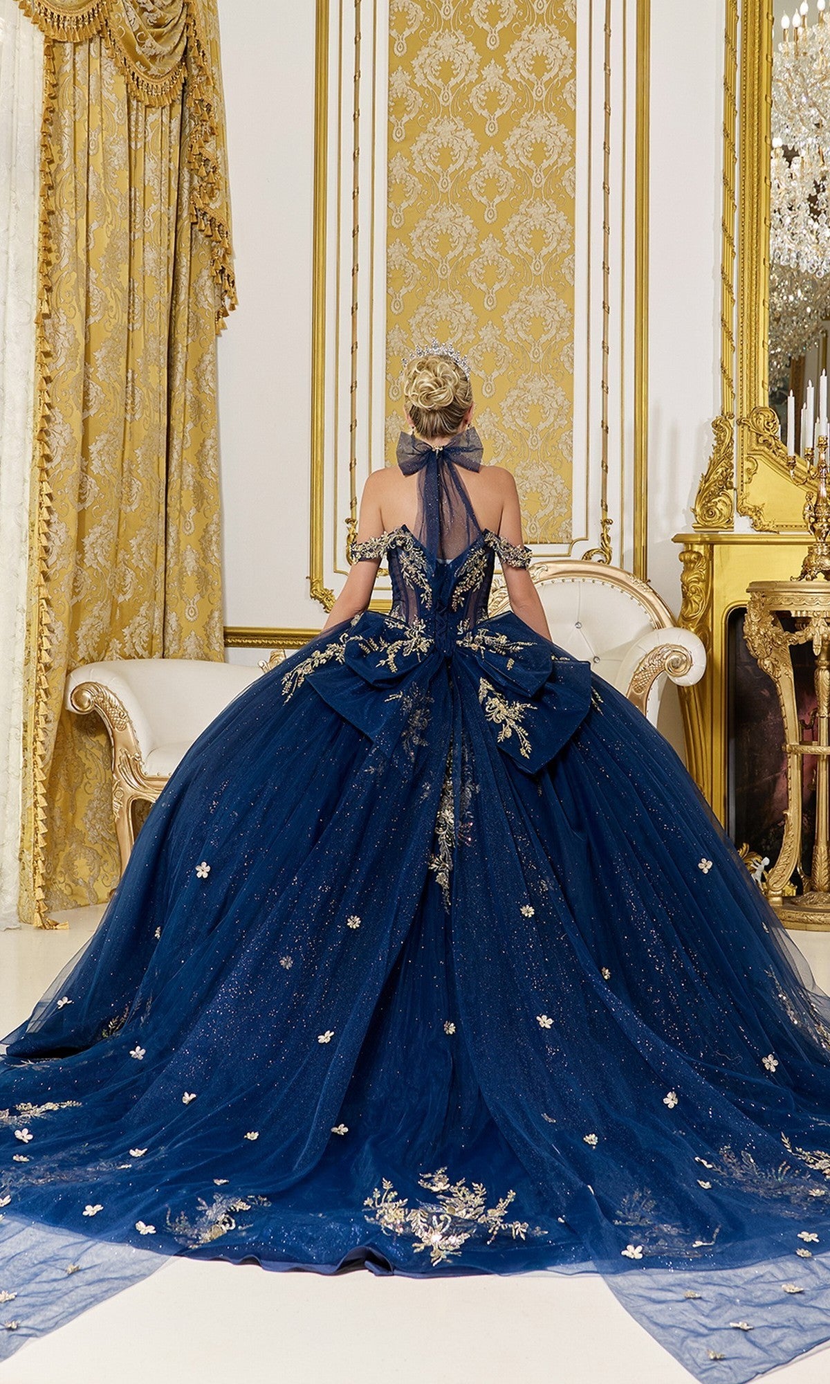 A woman in the Quinceanera Dress 5128 By Dancing Queen, a glitter tulle ball gown with gold applique, sits on a white and gold sofa in an opulent, gold-accented room.