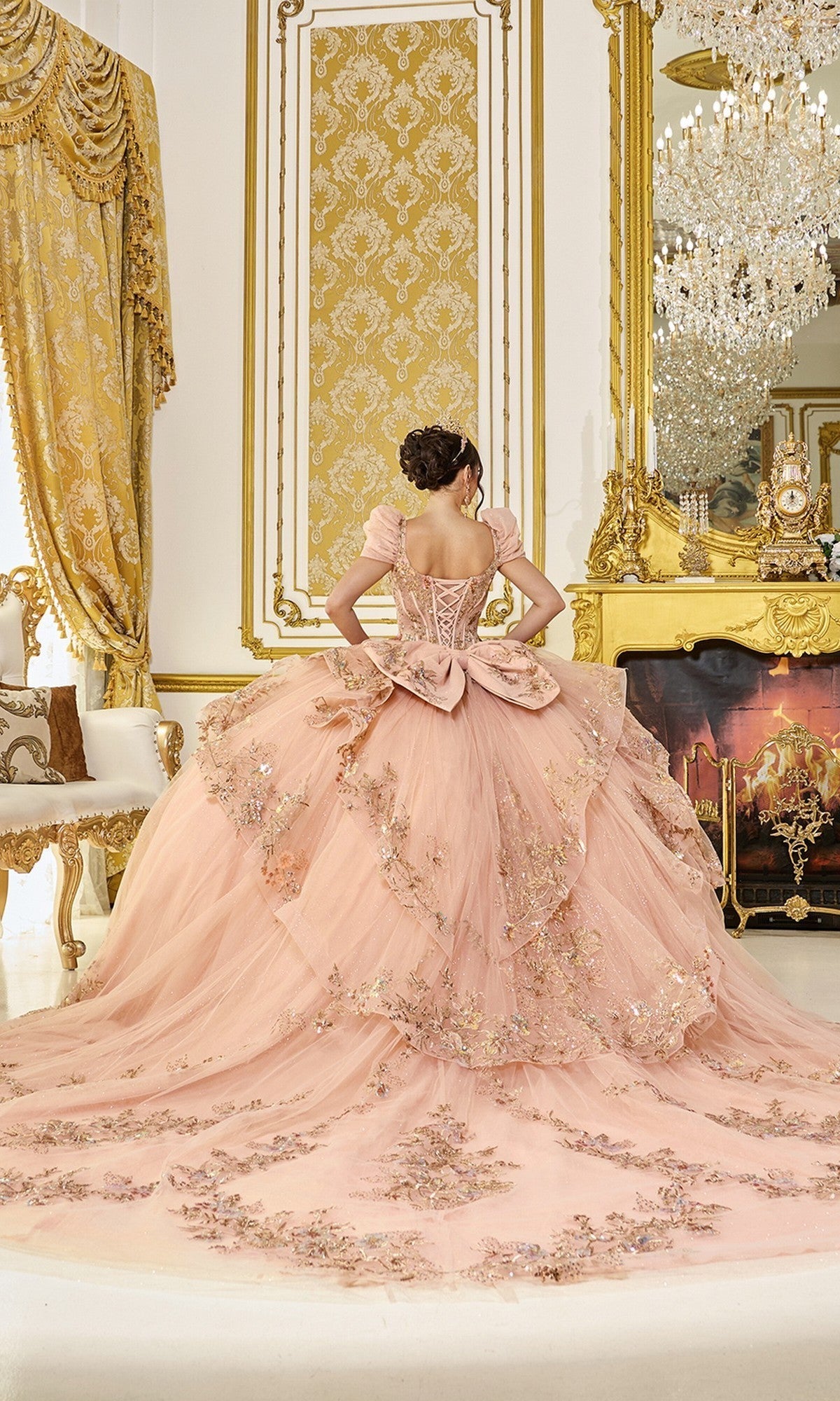 Wearing the Quinceanera Dress 5130 By Dancing Queen, a person stands with their back to the camera in an opulent, gold-accented room featuring a large mirror and a sparkling chandelier. The pink gown is adorned with 3D floral appliques.
