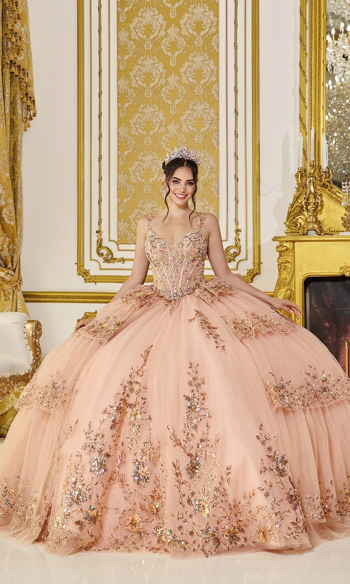 A young woman stands smiling at the camera in an ornate, gold-accented room, wearing the Quinceanera Dress 5131 by Dancing Queen in rose gold. The gown features a corset bodice and a flowing tulle ballgown skirt, with her tiara sparkling above the elaborately beaded design.