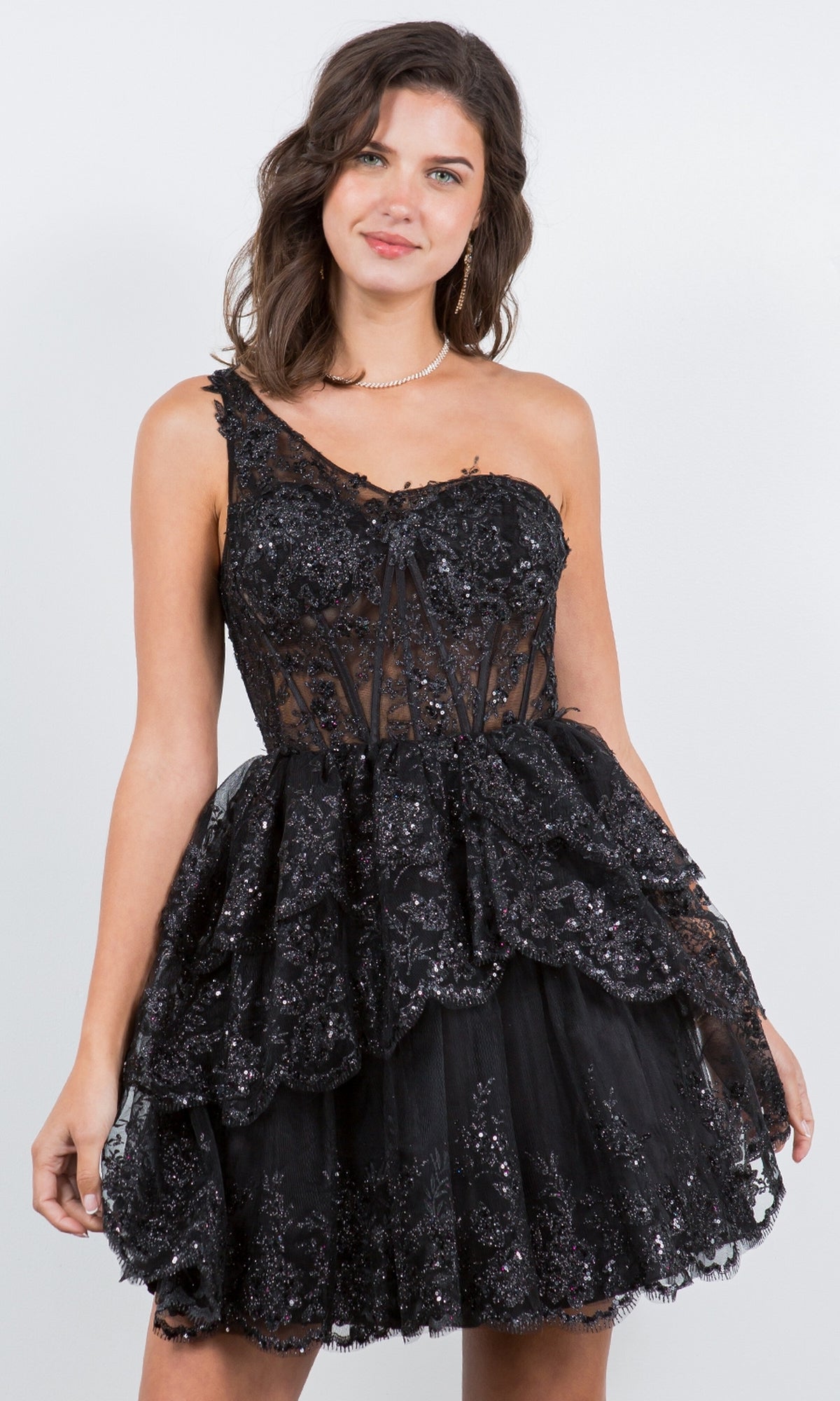 A woman stands against a plain background wearing the One-Shoulder Short Tiered Homecoming Dress 5132J, featuring black sparkly lace, a fitted bodice, and layered details.