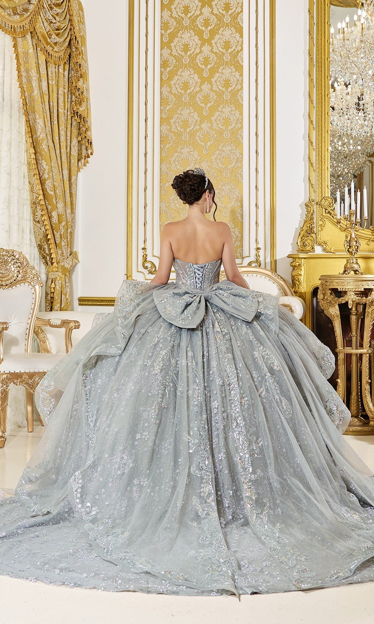 A woman wearing the Quinceanera Dress 5133 by Dancing Queen, featuring a shimmering finish, beaded corset bodice, and a large bow, stands with her back to the camera in an ornate room with gold accents.