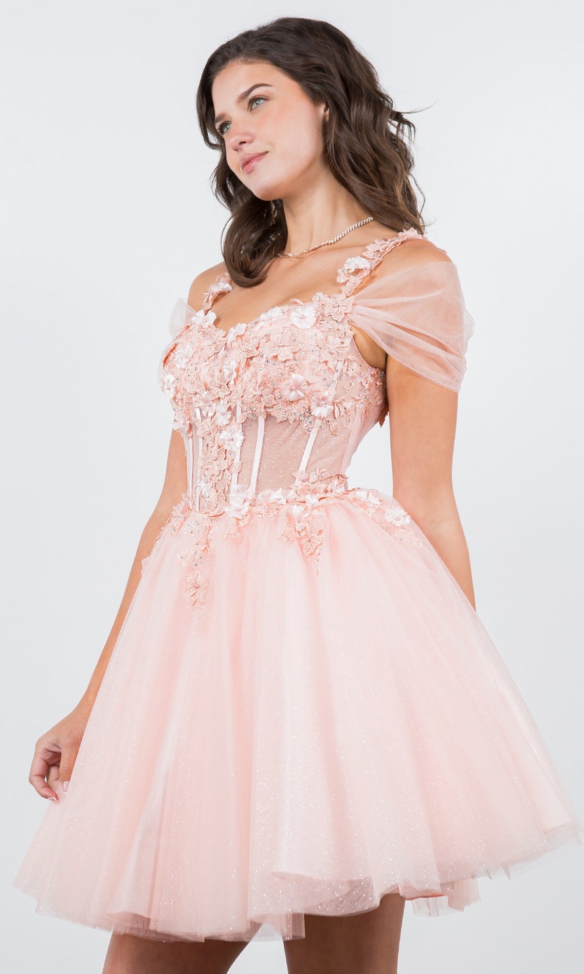 A woman poses against a plain background in the Embellished Short Glitter-Tulle Homecoming Dress, featuring light pink color, floral accents, sheer off-shoulder sleeves, and a short, sparkling tulle skirt.