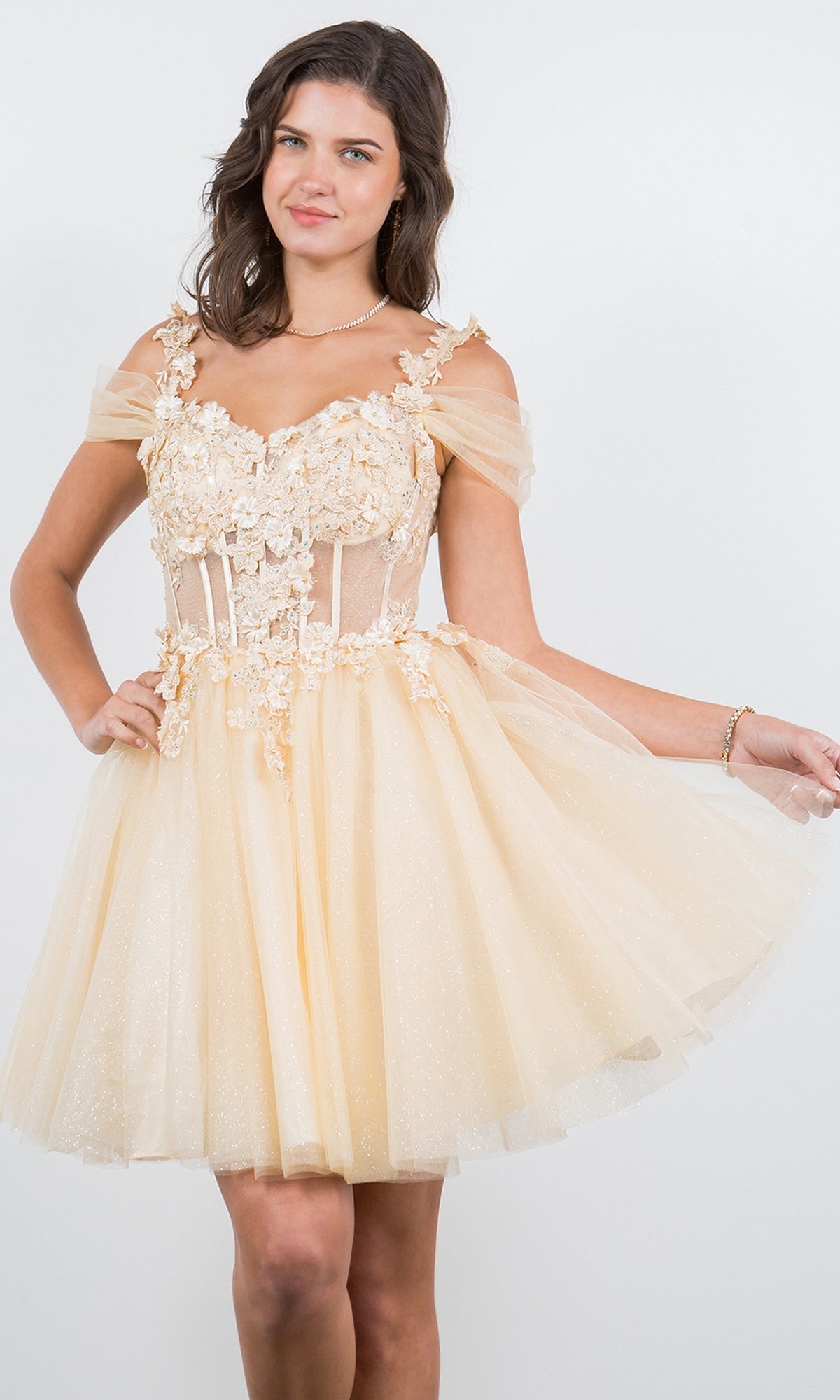 A young woman poses against a plain white background wearing the Embellished Short Glitter-Tulle Homecoming Dress, featuring an off-the-shoulder neckline, fitted bodice, and floral appliqué in light yellow.
