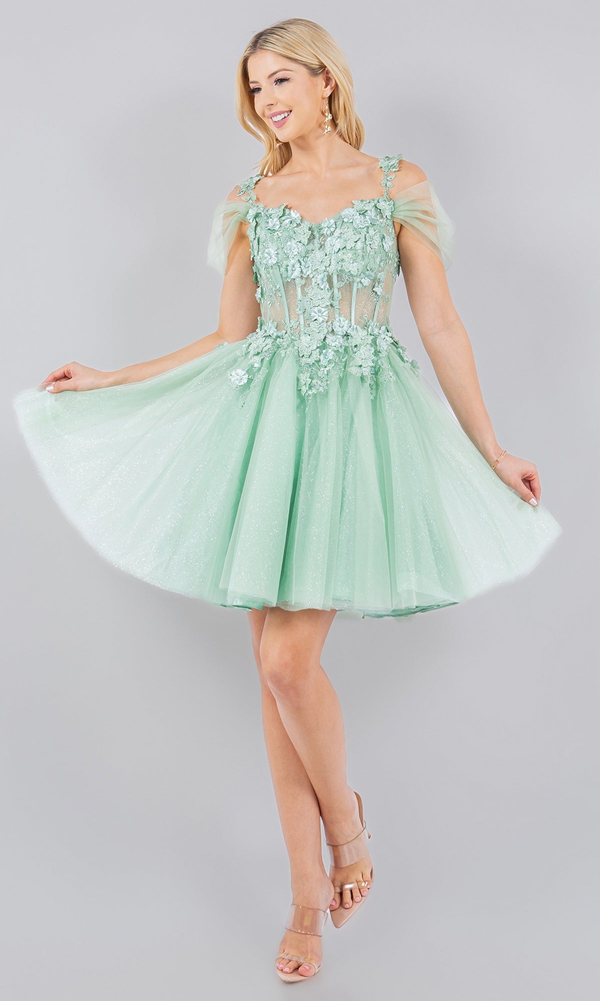 Woman modeling the Embellished Short Glitter-Tulle Homecoming Dress, a light green off-shoulder style with sheer floral details, against a plain light gray background.