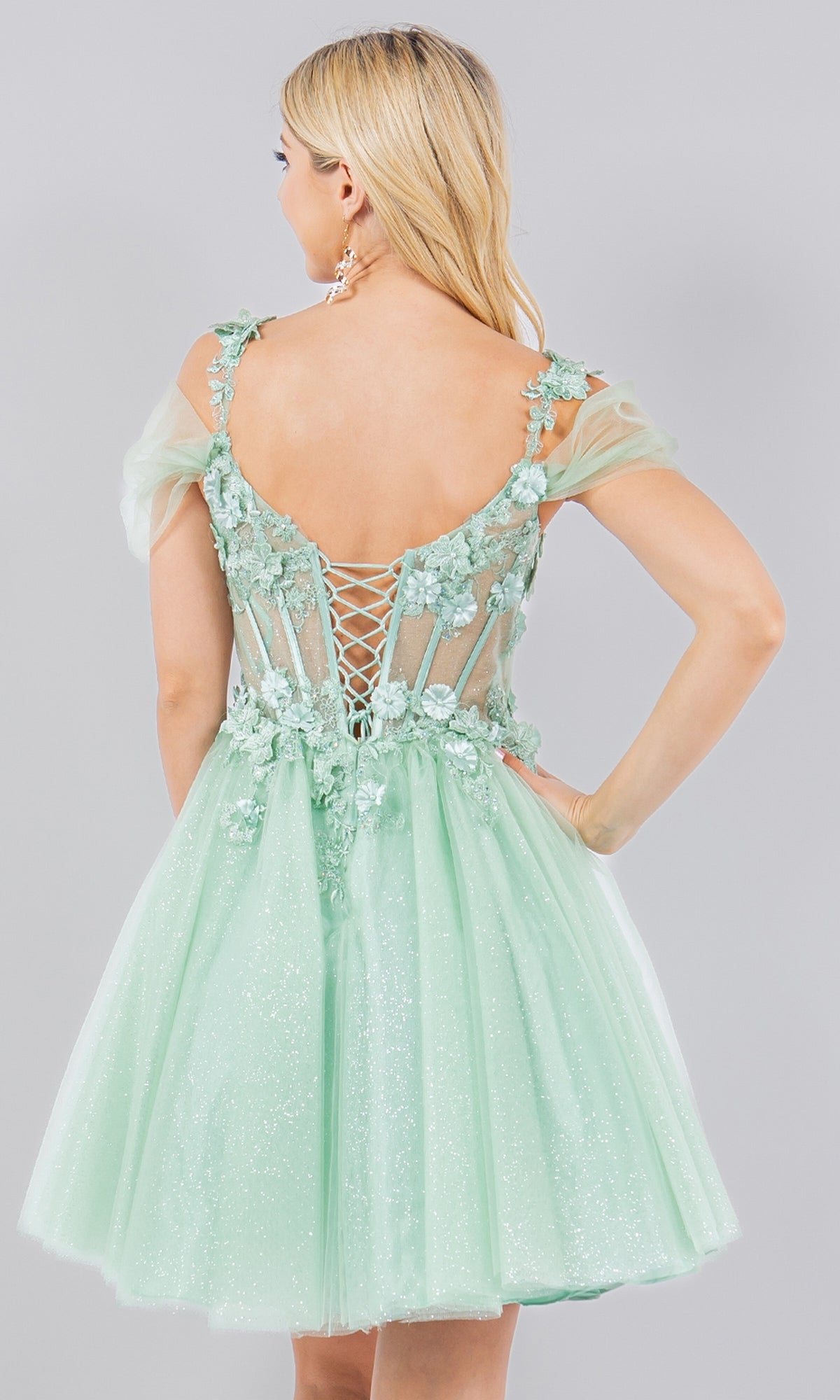 A woman in the Embellished Short Glitter-Tulle Homecoming Dress, featuring light green floral accents and a lace-up back, stands with her back to the camera.