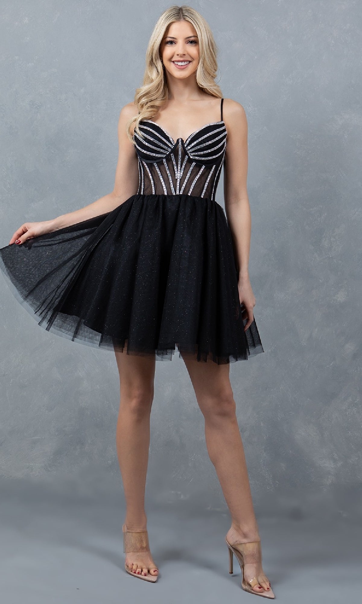 A woman poses against a gray textured background wearing the Cinderella Couture 5138J—a short black hoco dress featuring a glittery, fitted striped bodice and a flared tulle skirt.